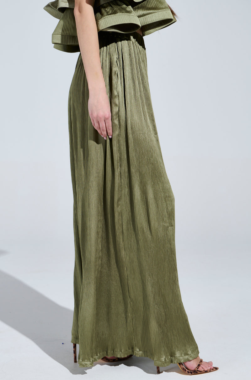 A person in FLY AWAY WITH ME PLEATED PALAZZO PANT and a matching top with ruffled sleeves stands sideways, their hand elegantly brushing the side. The plain floor and light gray background highlight the outfit's charm and hint at a wide-leg silhouette.
