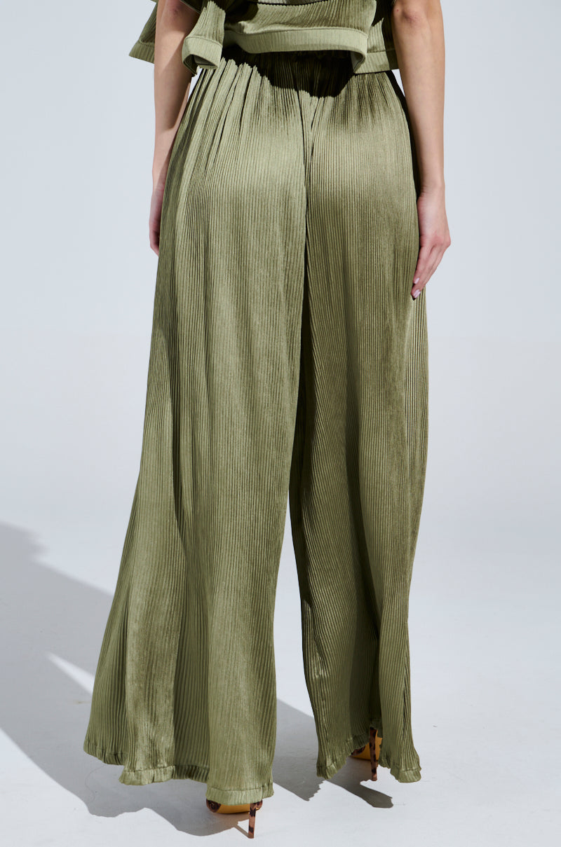 A person stands against a neutral background wearing the FLY AWAY WITH ME PLEATED PALAZZO PANT in olive green. Their hands rest on their sides, and tan pointed heels complement the flowy, wide-leg silhouette, adding a soft sheen to the satin fabric.