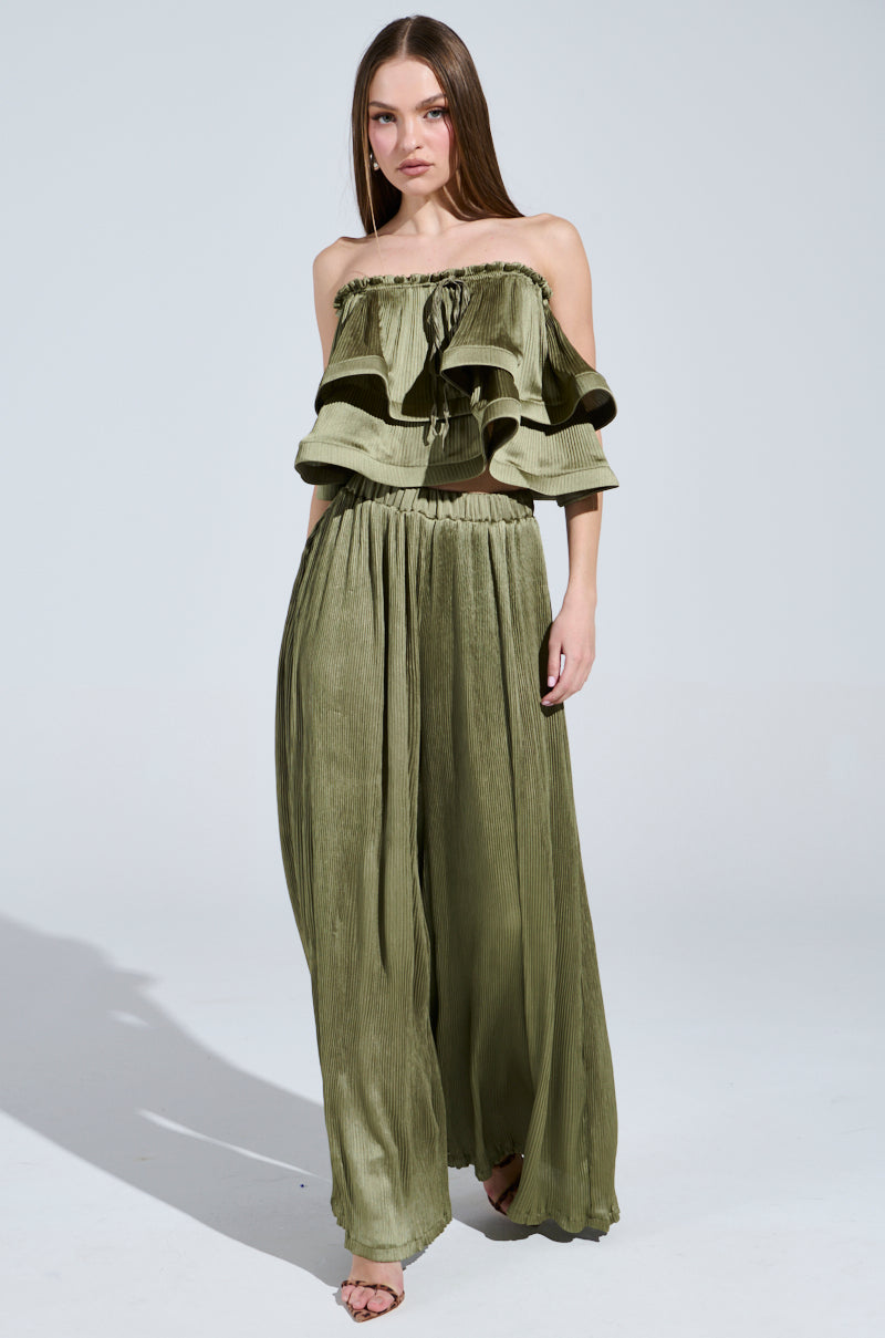 A woman gracefully stands barefoot, wearing an olive green off-shoulder ruffled top with elegant "Fly Away With Me Pleated Palazzo Pants." Her straight hair falls loosely around her against a plain, light background.