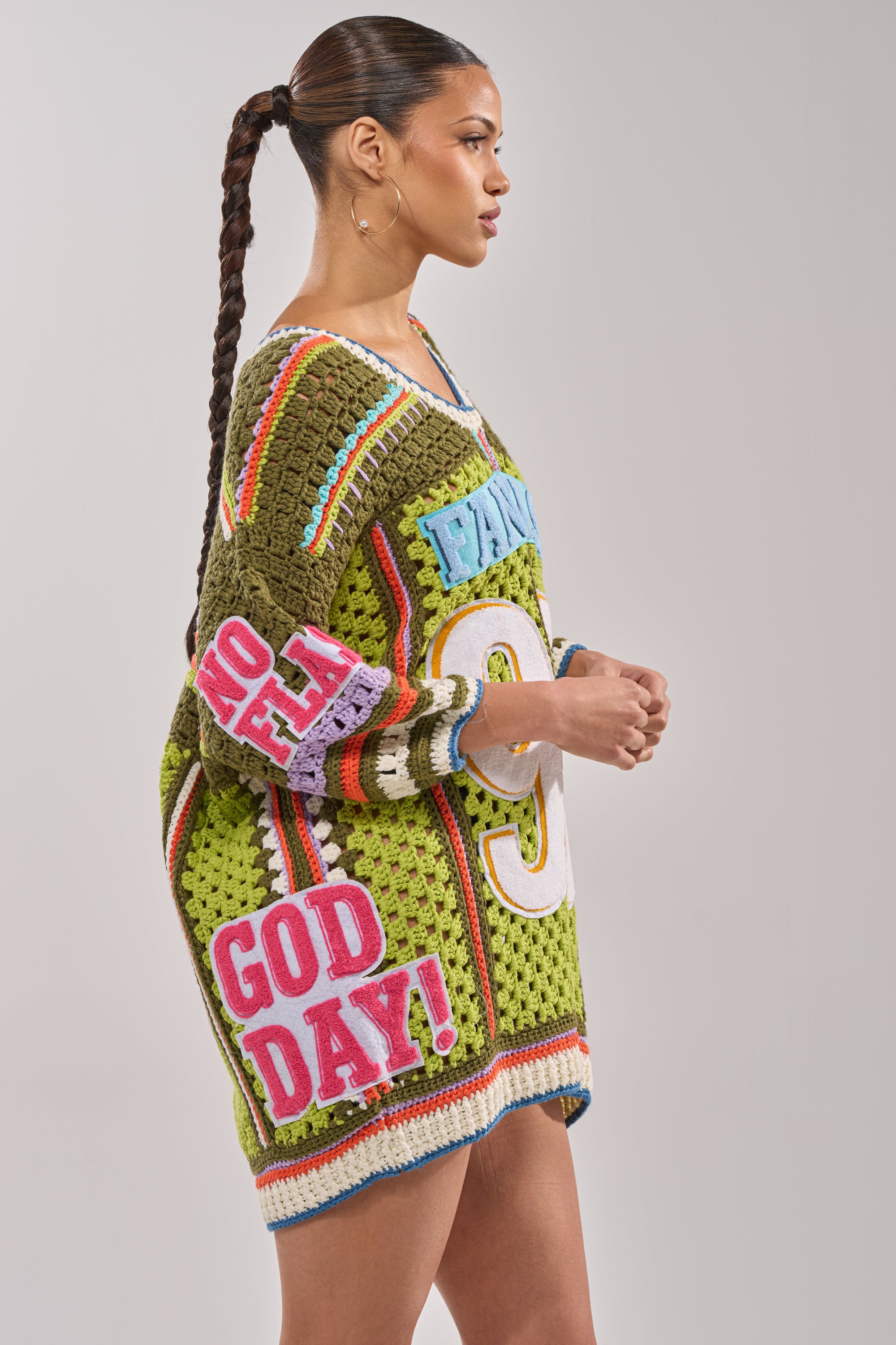 A woman with a sleek ponytail models the YESSSS CROCHET SWEATER, an oversized, colorful piece with bold text patches and varsity-inspired granny-square crochet details, set against a plain background. 6