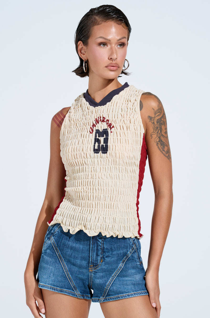 A woman with short dark hair and hoop earrings wears the cream-colored ADDICTED RUCHED JERSEY TOP, denim shorts, and shows a visible arm tattoo while standing against a light background.