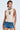 A woman with short dark hair and hoop earrings wears the cream-colored ADDICTED RUCHED JERSEY TOP, denim shorts, and shows a visible arm tattoo while standing against a light background.
