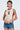 A woman with short, slicked-back hair, visible arm tattoo, and denim shorts wears the ADDICTED RUCHED JERSEY TOP, standing against a plain light background.