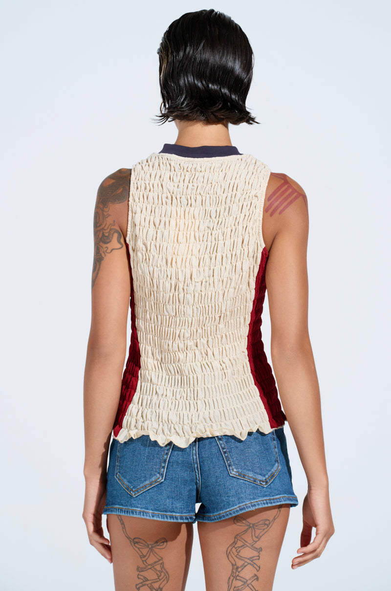 Person with short, wet-look dark hair wears the ADDICTED RUCHED JERSEY TOP in cream and red with denim shorts, seen from behind. Tattoos show on their arms and legs. Plain light background gives a sporty-chic vibe.