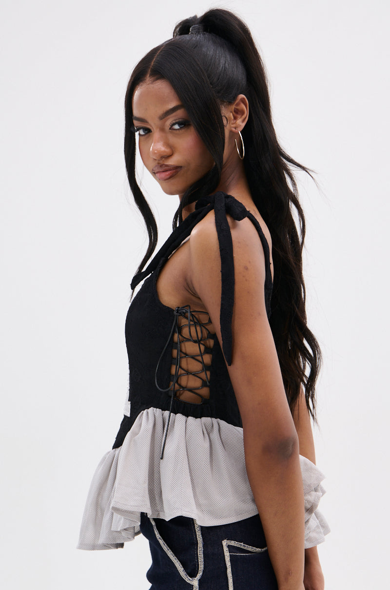 A woman with long, wavy black hair in a high ponytail wears the OFFICIAL PRINCESS LACE PEPLUM CORSET TOP and dark jeans with white stitching, posing sideways and looking at the camera.