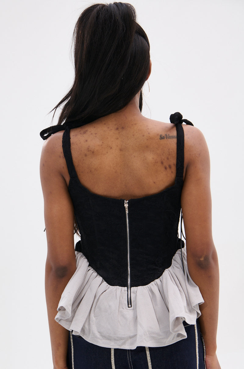 A woman with long dark hair faces away, showing her upper back tattoo while wearing the OFFICIAL PRINCESS LACE PEPLUM CORSET TOP—a black sleeveless piece with a zipper closure and mesh peplum hem—paired with dark pants.