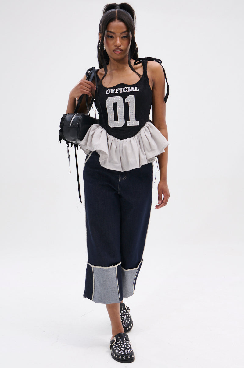 A woman poses in the OFFICIAL PRINCESS LACE PEPLUM CORSET TOP with "OFFICIAL 01," styled with wide-leg dark jeans featuring light cuffs, black studded slip-on shoes, and a small black shoulder bag.