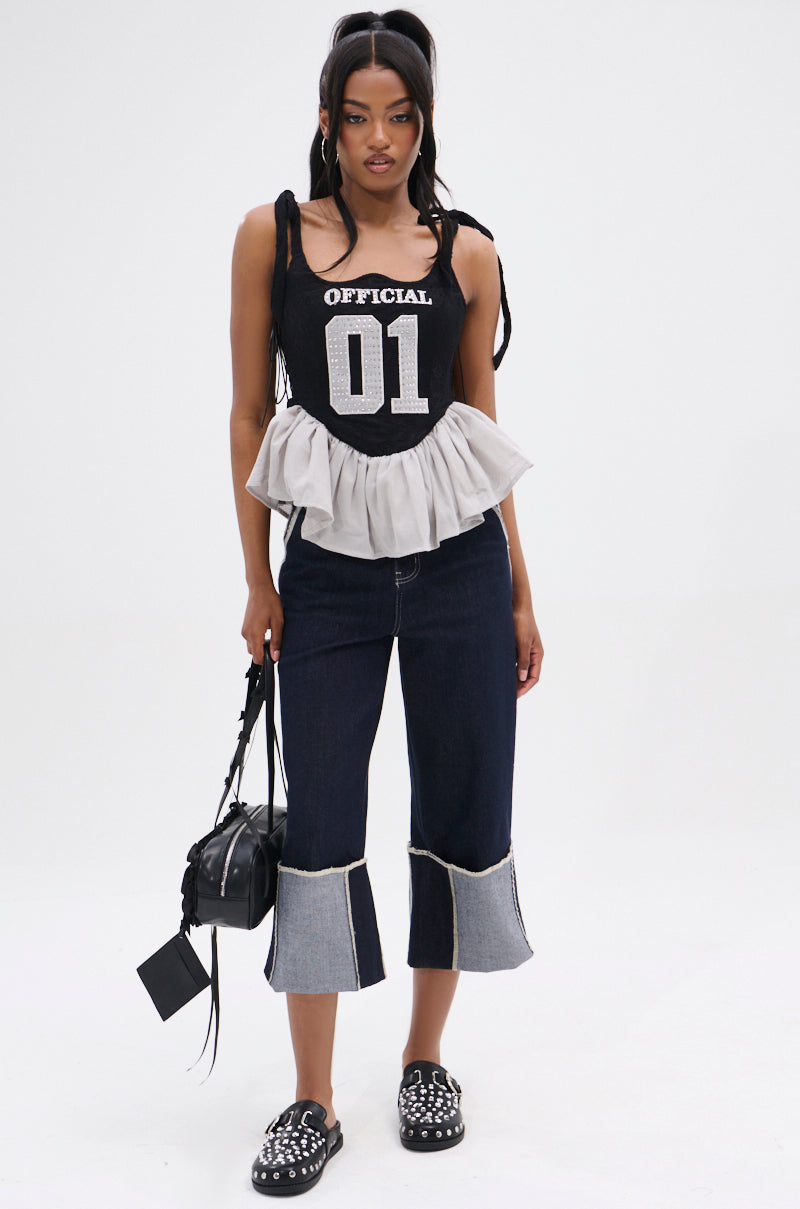 A woman stands facing forward, wearing the OFFICIAL PRINCESS LACE PEPLUM CORSET TOP, cropped two-tone denim pants, studded black slip-on shoes, and holding a small black handbag.