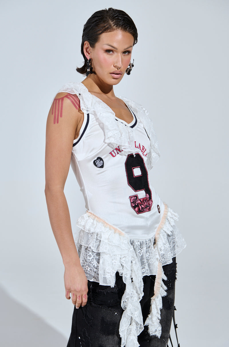 A person exudes a sporty-chic vibe, confidently posing in a white "PLAY WITH MY HEART RUFFLE JERSEY" with the number 9 and lace details. Paired with stretchy black pants, they showcase slicked-back hair, bold makeup, and statement earrings against a plain gray background.