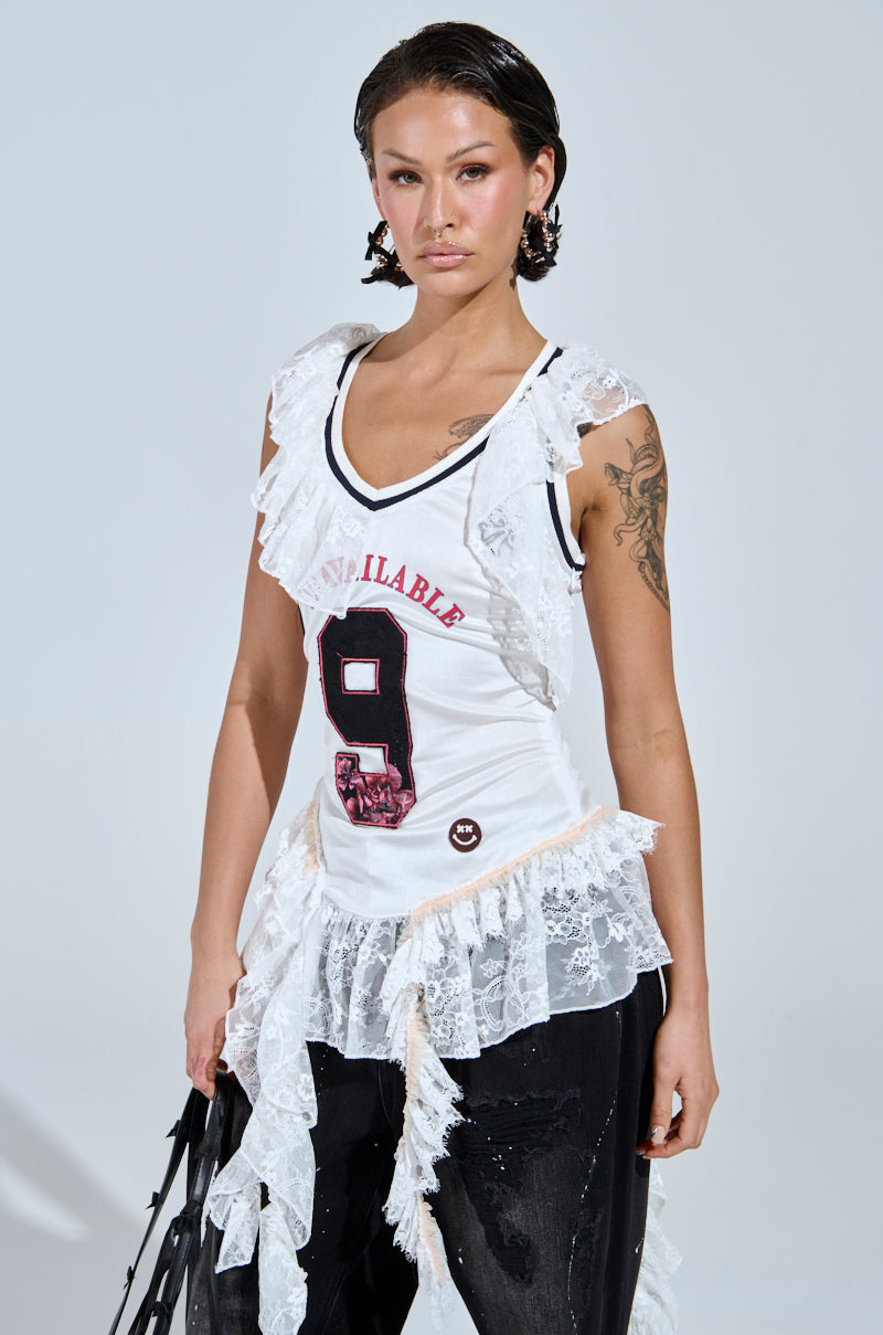 A person sports a chic ensemble, featuring the PLAY WITH MY HEART RUFFLE JERSEY in white with "9" and "Unavailable." Their wet-look hair and tattooed arm pair well with black lace-detail pants, while they carry a sleek black bag.