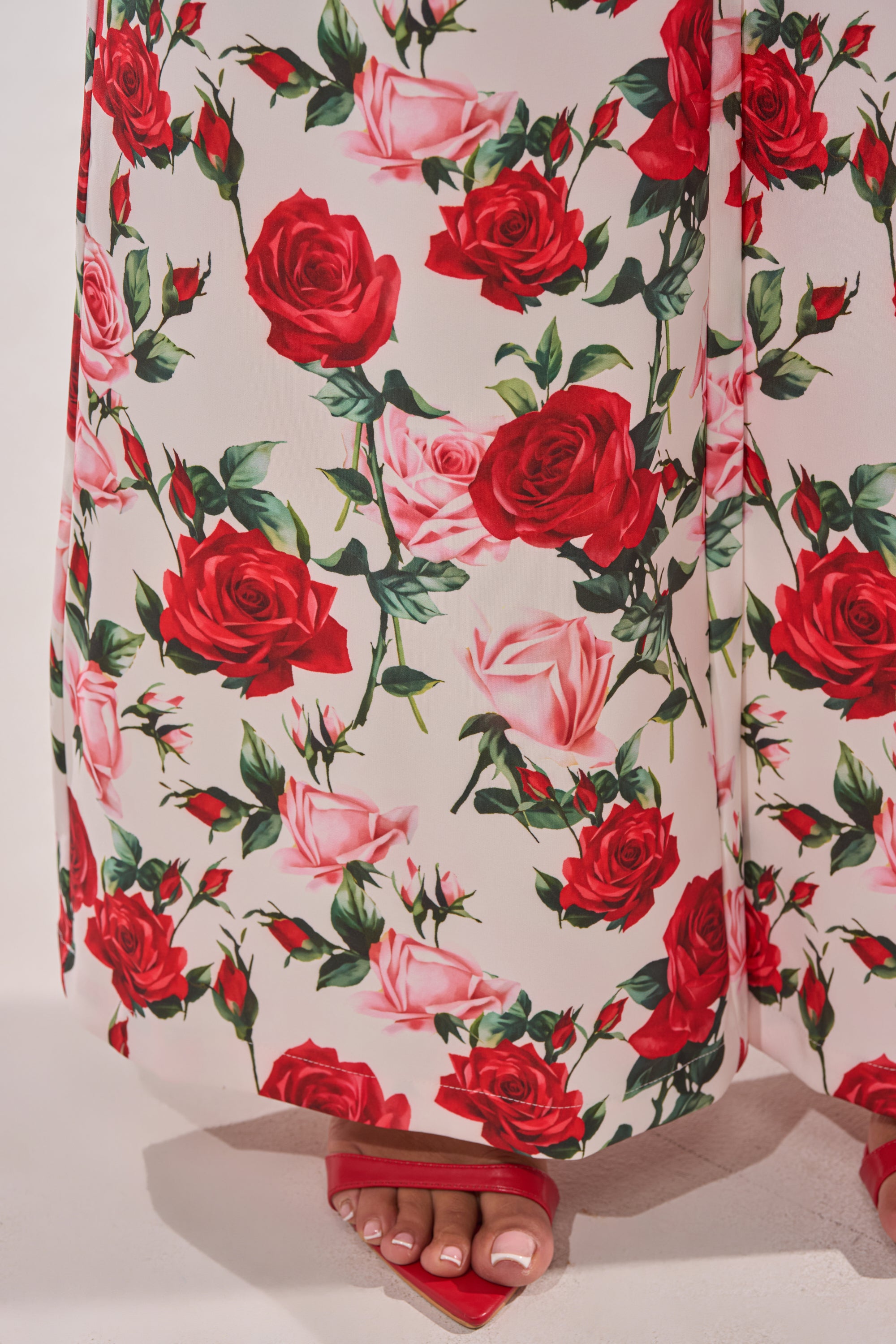 A close-up of the FULL BLOOM FLORAL TROUSER, featuring a red and pink rose pattern, with a person’s feet in red open-toed heeled sandals peeking from beneath the wide-leg hem. 9