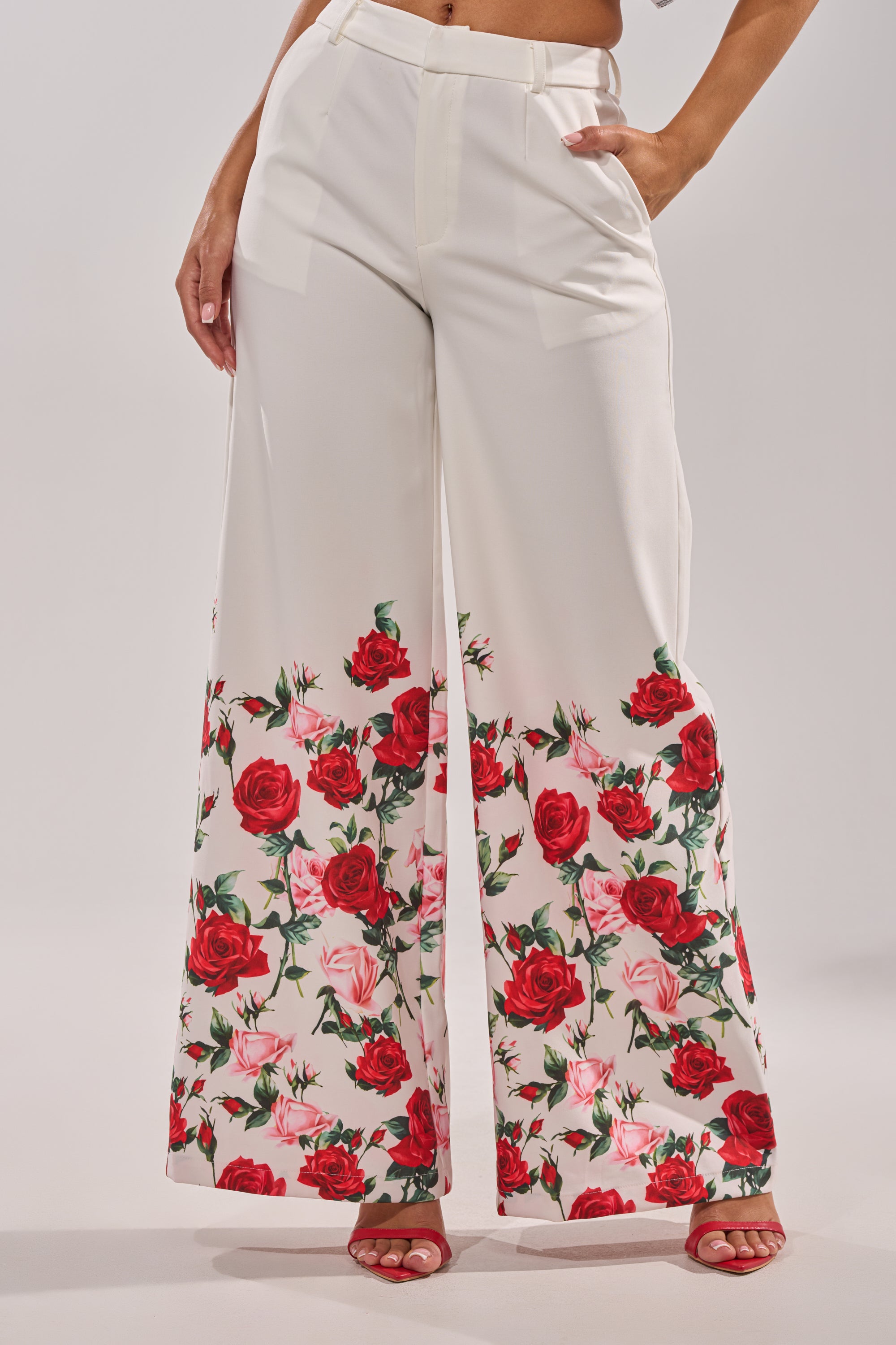 A person wears the FULL BLOOM FLORAL TROUSER, featuring high-rise waist and wide-leg design with a bold rose print near the hem, paired with red high-heeled sandals. The background is plain and light-colored. 1