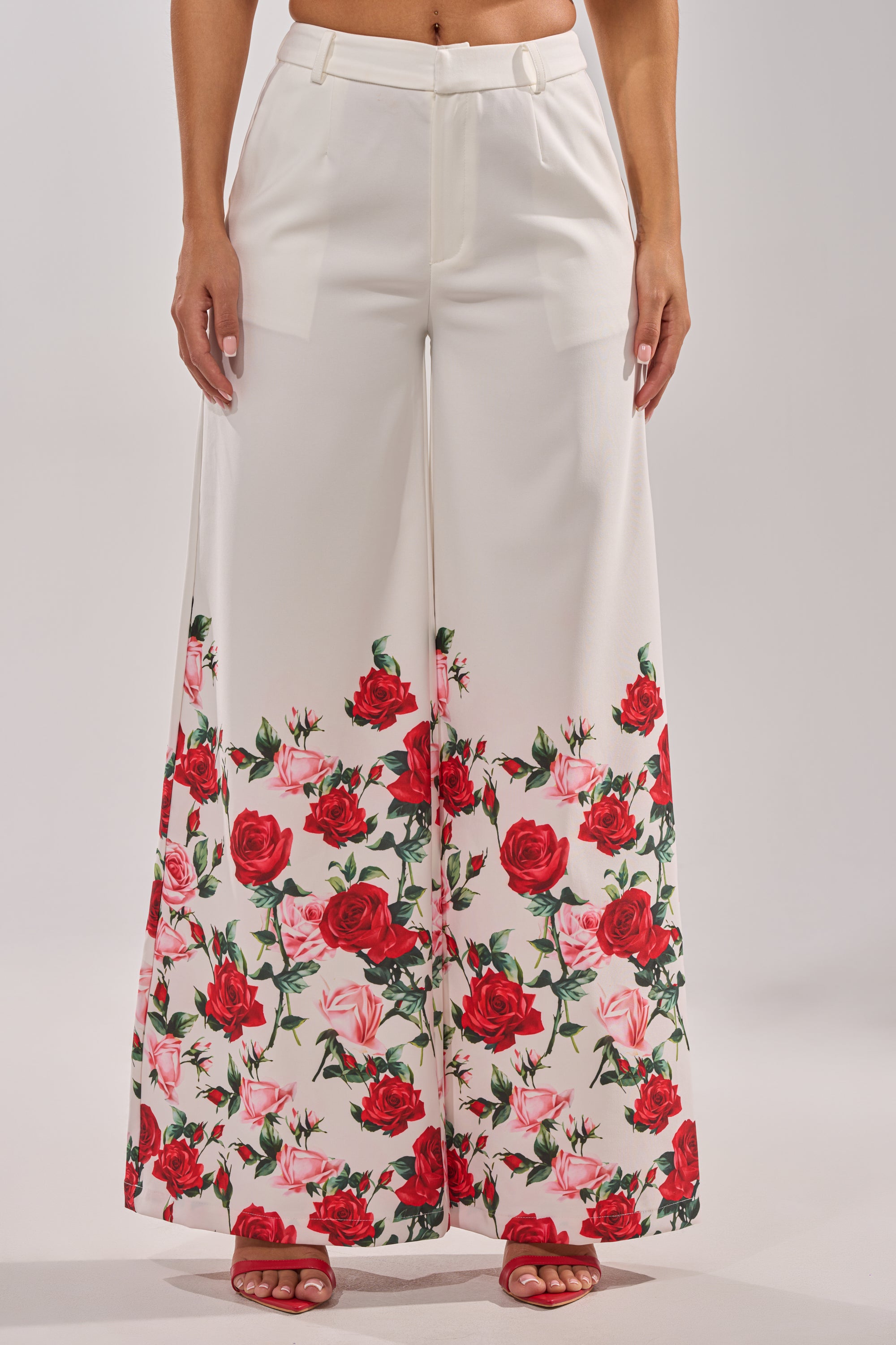 Someone wears the FULL BLOOM FLORAL TROUSER, a high-rise, wide-leg pant with bold red and pink rose prints on the lower half, styled with red open-toe heels against a plain light background. 2