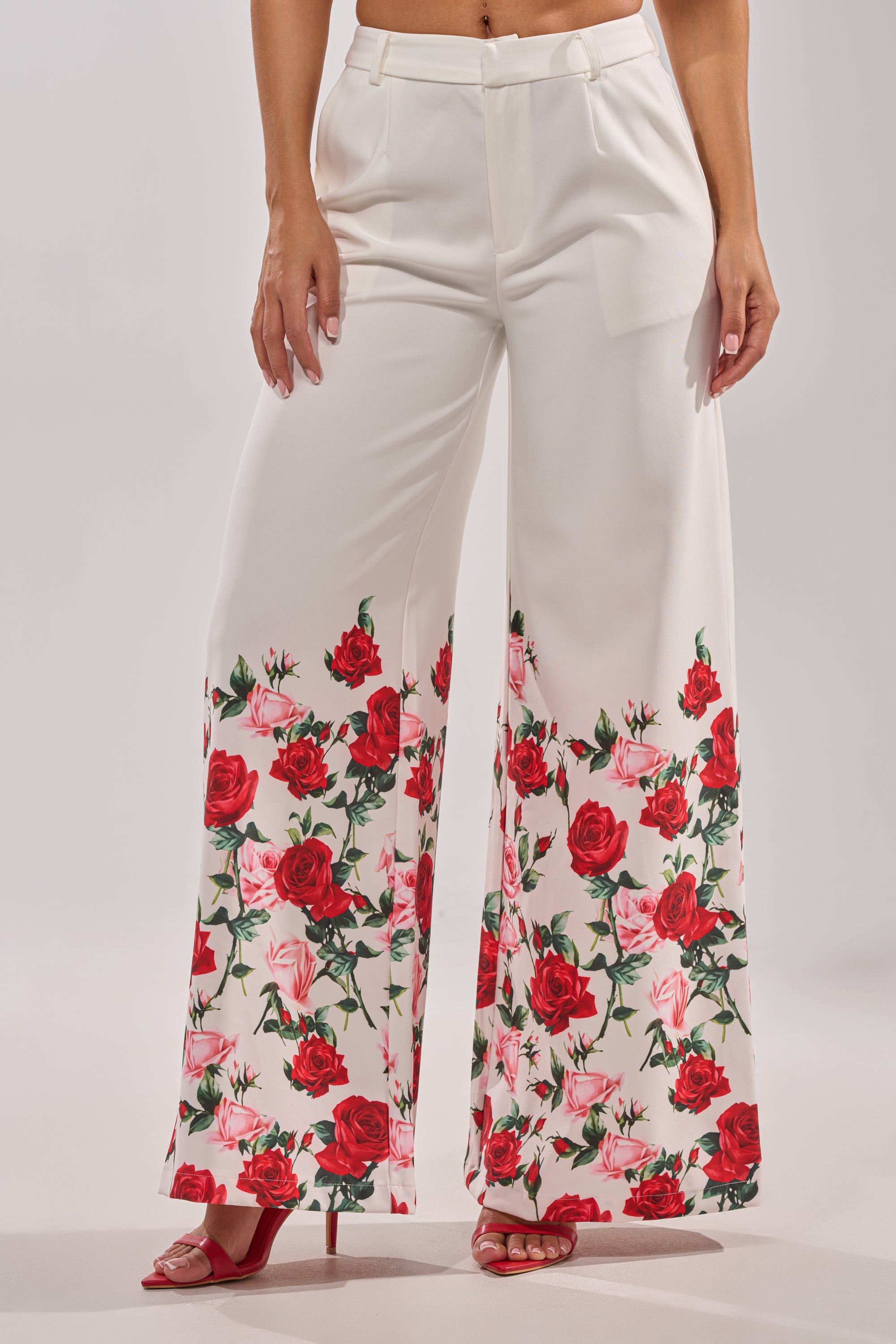 Wearing the FULL BLOOM FLORAL TROUSER—wide-leg, high-rise pants with a red and pink rose print near the hem—paired elegantly with red high-heeled sandals. 3