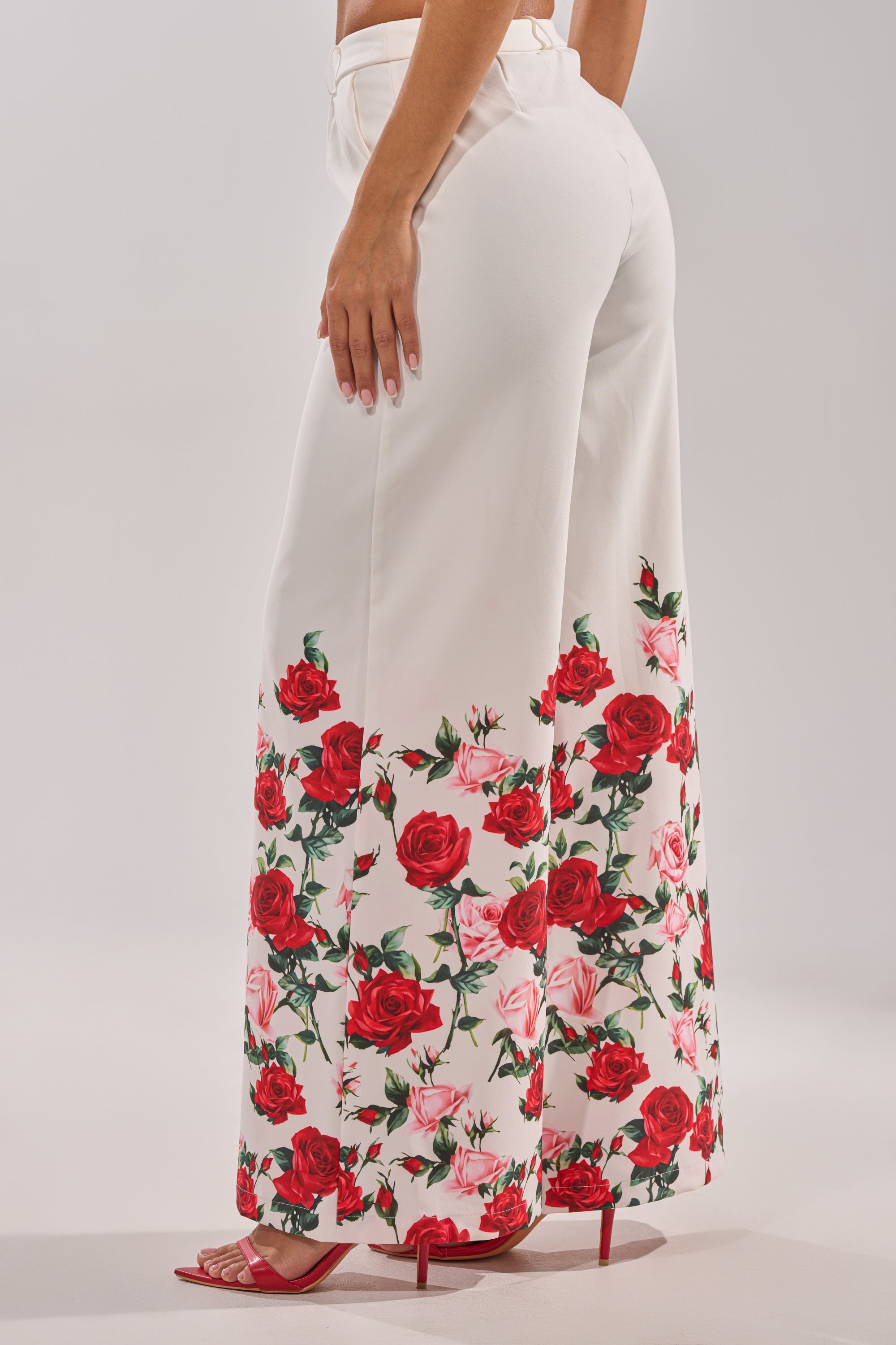 A woman models the FULL BLOOM FLORAL TROUSER with a high-rise waist and wide-leg fit, featuring red and pink rose prints on the lower legs. She pairs them with red heeled sandals in a side and back view. 4