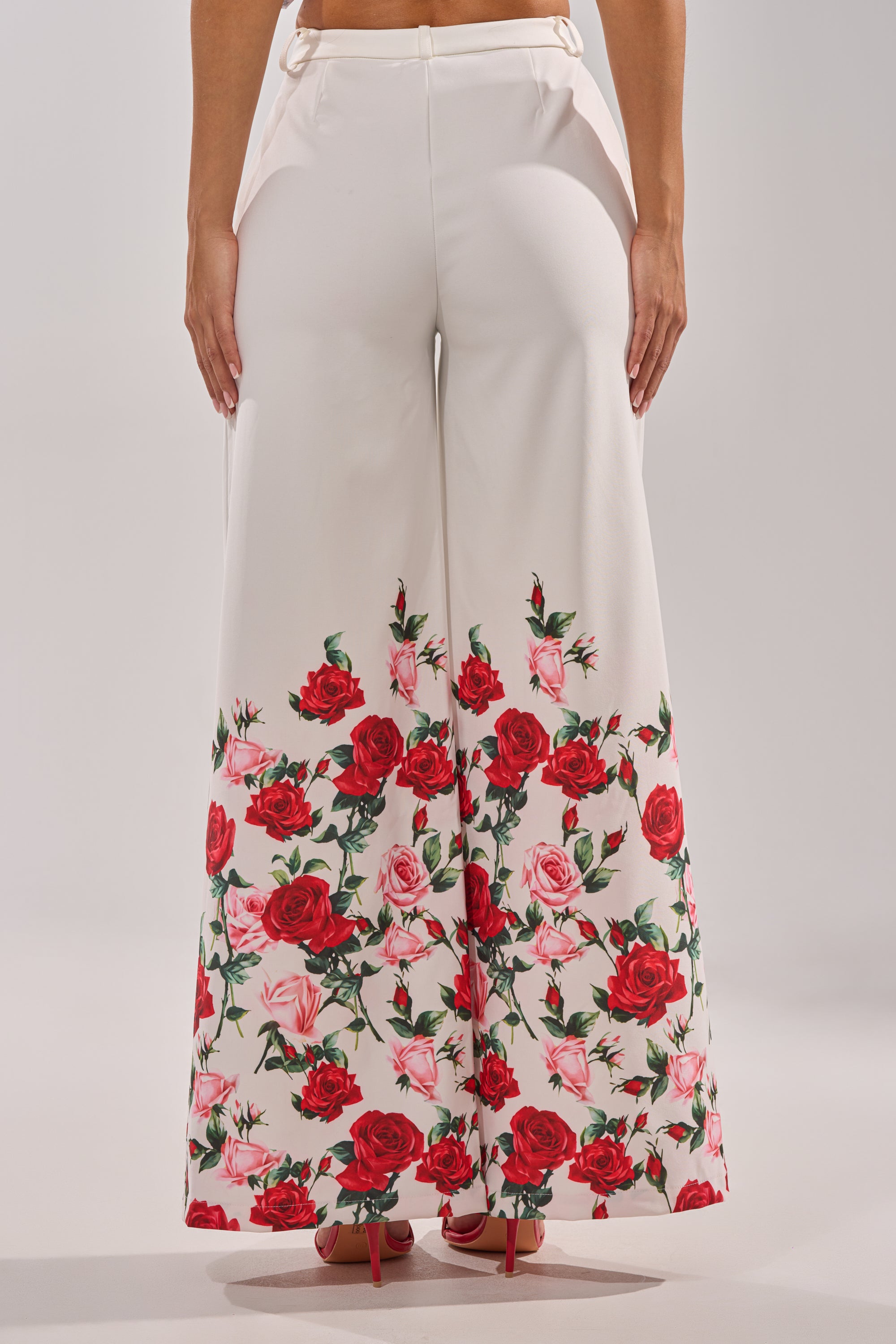 Someone stands against a plain background wearing the FULL BLOOM FLORAL TROUSER, featuring a high-rise waist and wide-leg fit with red and pink rose prints at the hem, paired with red high-heeled shoes. 5