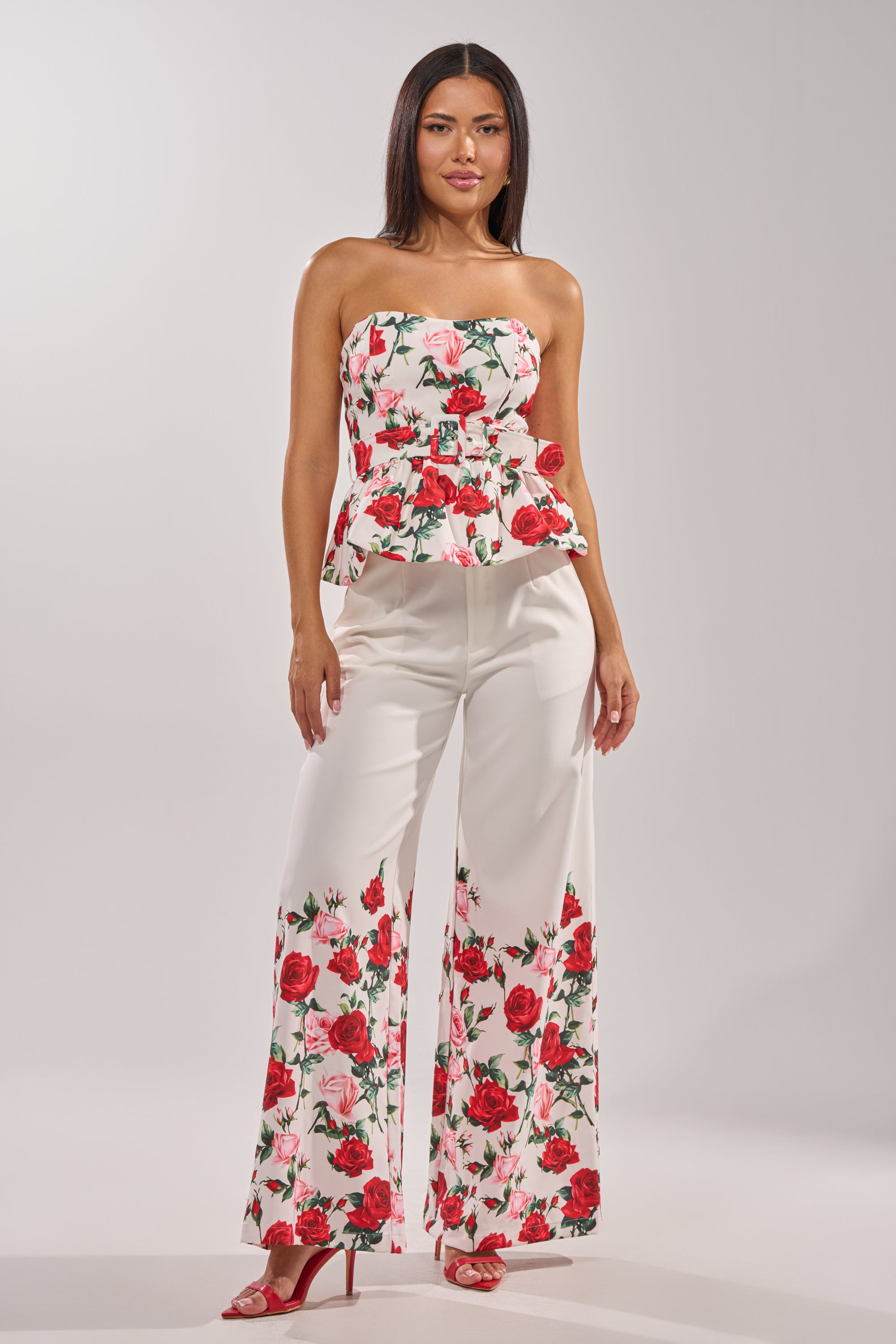 A woman poses against a plain light background in the FULL BLOOM FLORAL TROUSER, featuring white high-rise wide-leg pants with red rose prints, paired with a matching strapless peplum top and red high-heeled sandals. 6