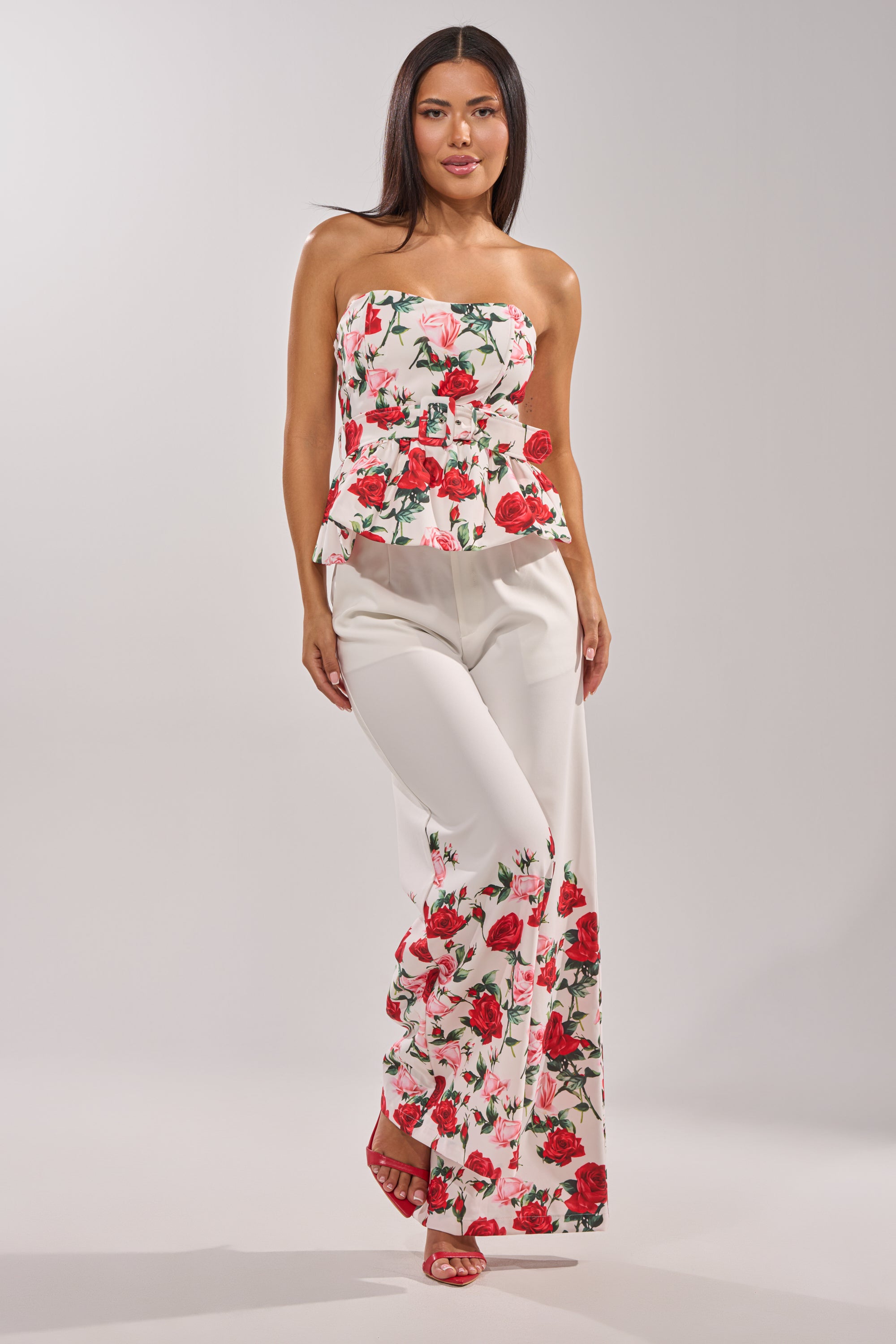 A woman poses in the FULL BLOOM FLORAL TROUSER, featuring a high-rise waist and rose print, paired with a matching strapless white floral top and red open-toe heels against a plain light background. 7