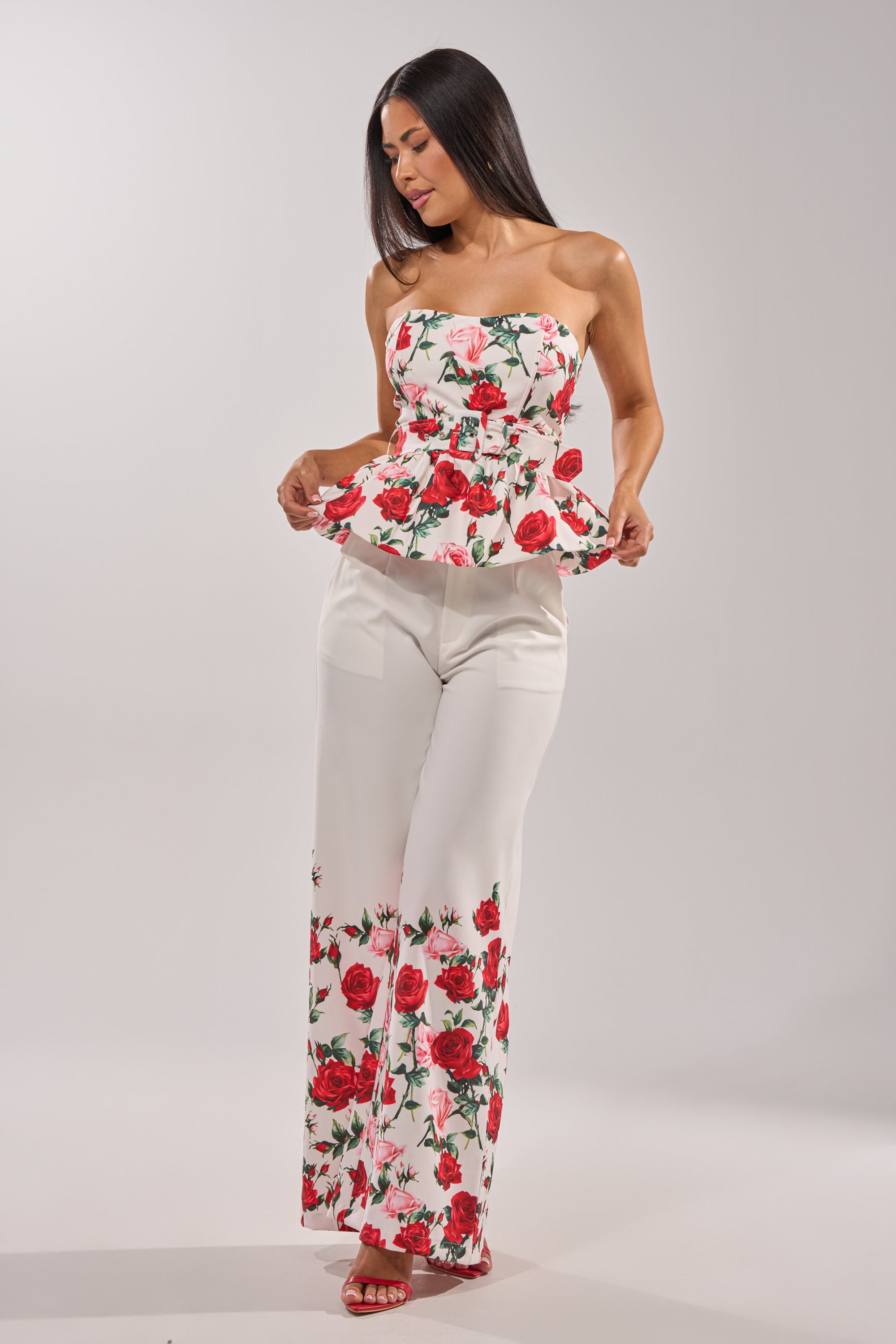 A woman poses against a plain light background, looking down and holding the edges of her strapless white peplum top while wearing the FULL BLOOM FLORAL TROUSER—high-rise, wide-leg pants with a rose print. 8