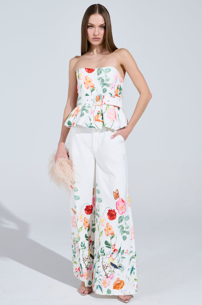 A woman confidently wears the IRIS FLORAL TROUSER set, with a strapless peplum top and high-rise wide-leg pants. She holds a pink feathered clutch in one hand while the other rests in her pocket, adorned with colorful flowers and butterflies.