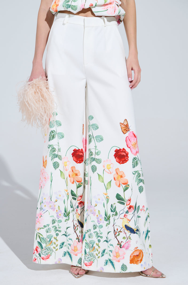 Someone in IRIS FLORAL TROUSER, known for its wide-leg silhouette and high-rise colorful bird and flower prints, stands against a light background with a fluffy pink accessory.