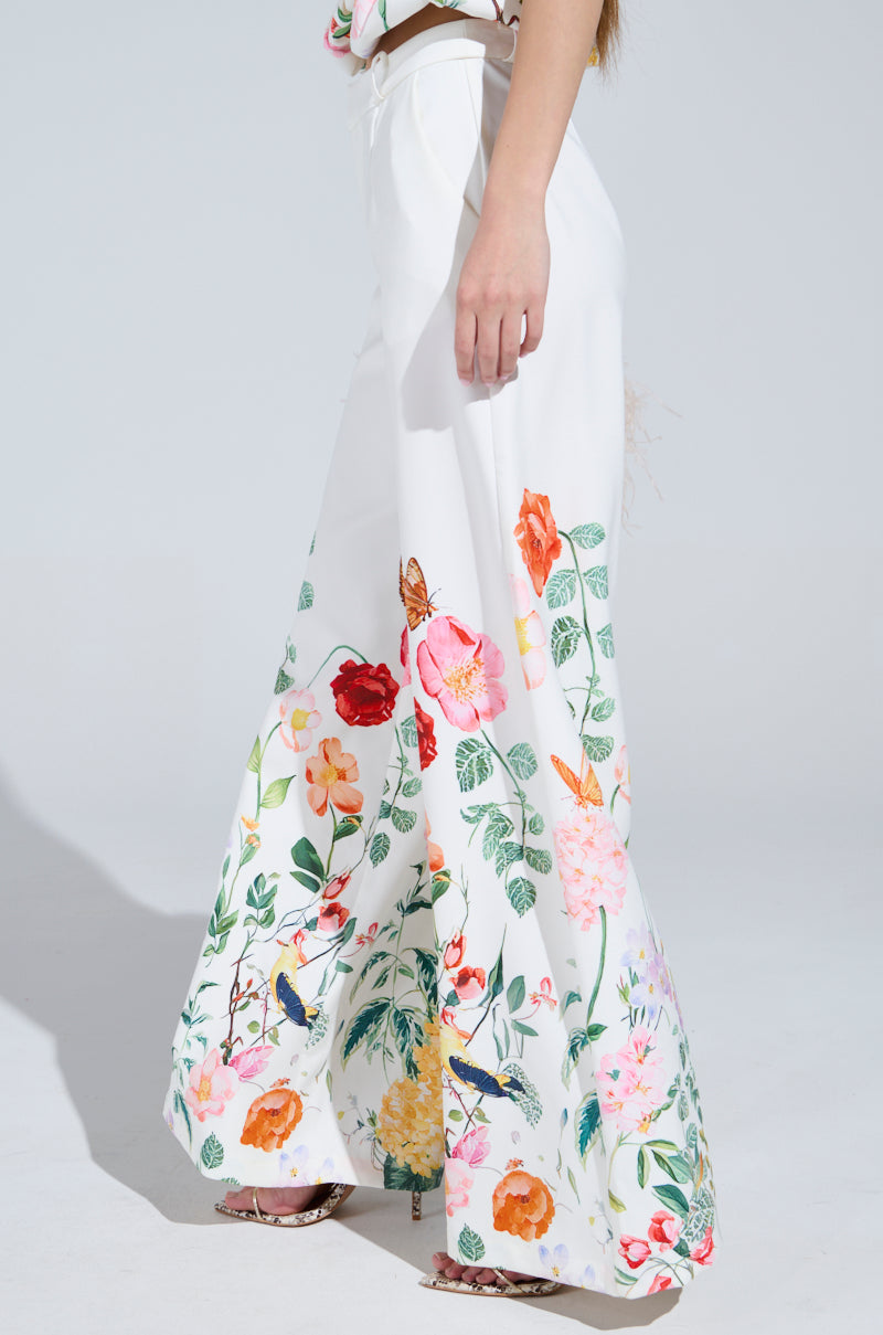 A woman models the IRIS FLORAL TROUSER, boasting a wide-leg silhouette with vibrant floral prints near the hem. The high-rise fit enhances her visible top as she elegantly poses against a plain background in chic open-toed shoes.
