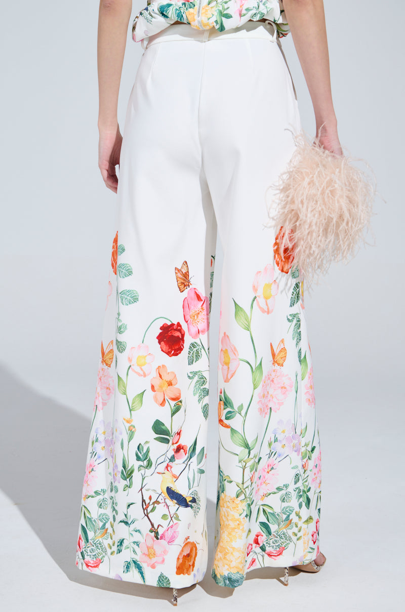 A person dons high-waisted IRIS FLORAL TROUSER with a wide-leg design and vibrant florals, clutching a beige feathered bag. The intricate patterns are showcased from behind on a plain light gray backdrop.