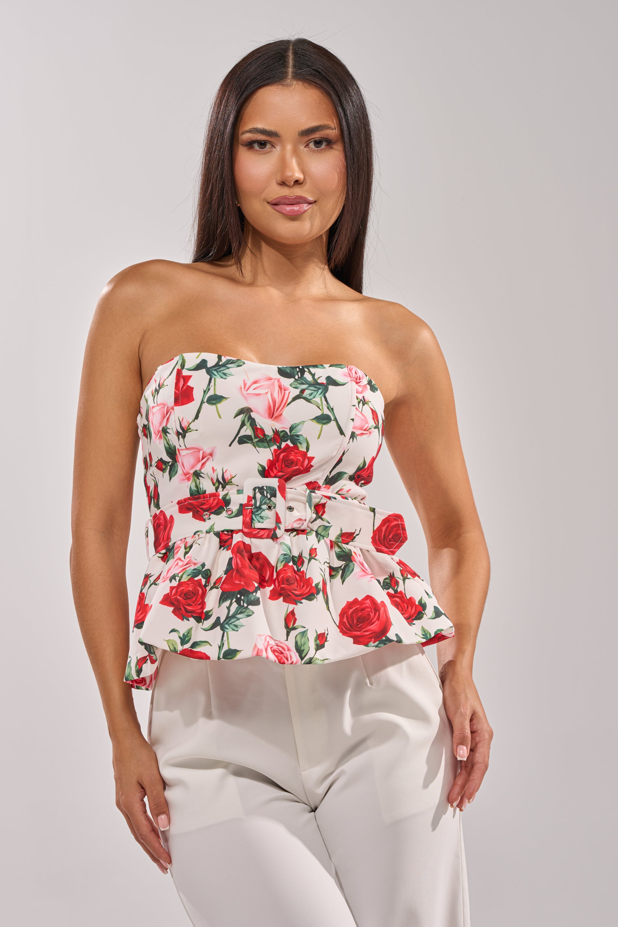 A woman with long dark hair models the FULL BLOOM FLORAL CORSET, a strapless top with red and pink rose print, paired with white pants against a plain light background. 0