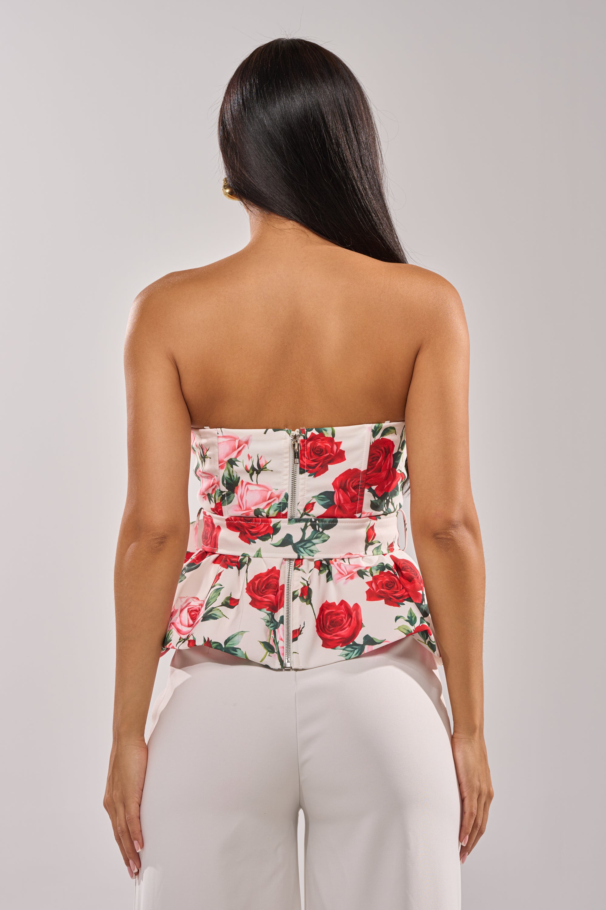 A woman with long dark hair, seen from behind, wears the FULL BLOOM FLORAL CORSET—a white strapless top with a red rose pattern—paired with white high-waisted pants against a plain light background. 3