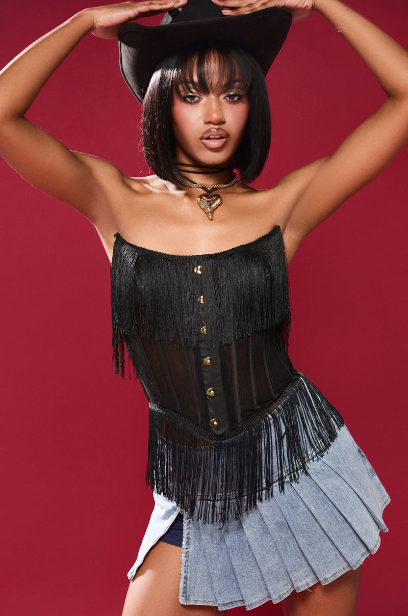 In a bold red backdrop, a figure radiates confidence in a FLAPPER GIRL CORSET TOP, black hat, and denim mini skirt. Their mid-length hair complements the vintage-chic look, accentuated by a necklace. The ensemble blends western and modern styles with structural boning for enhanced fashion appeal.