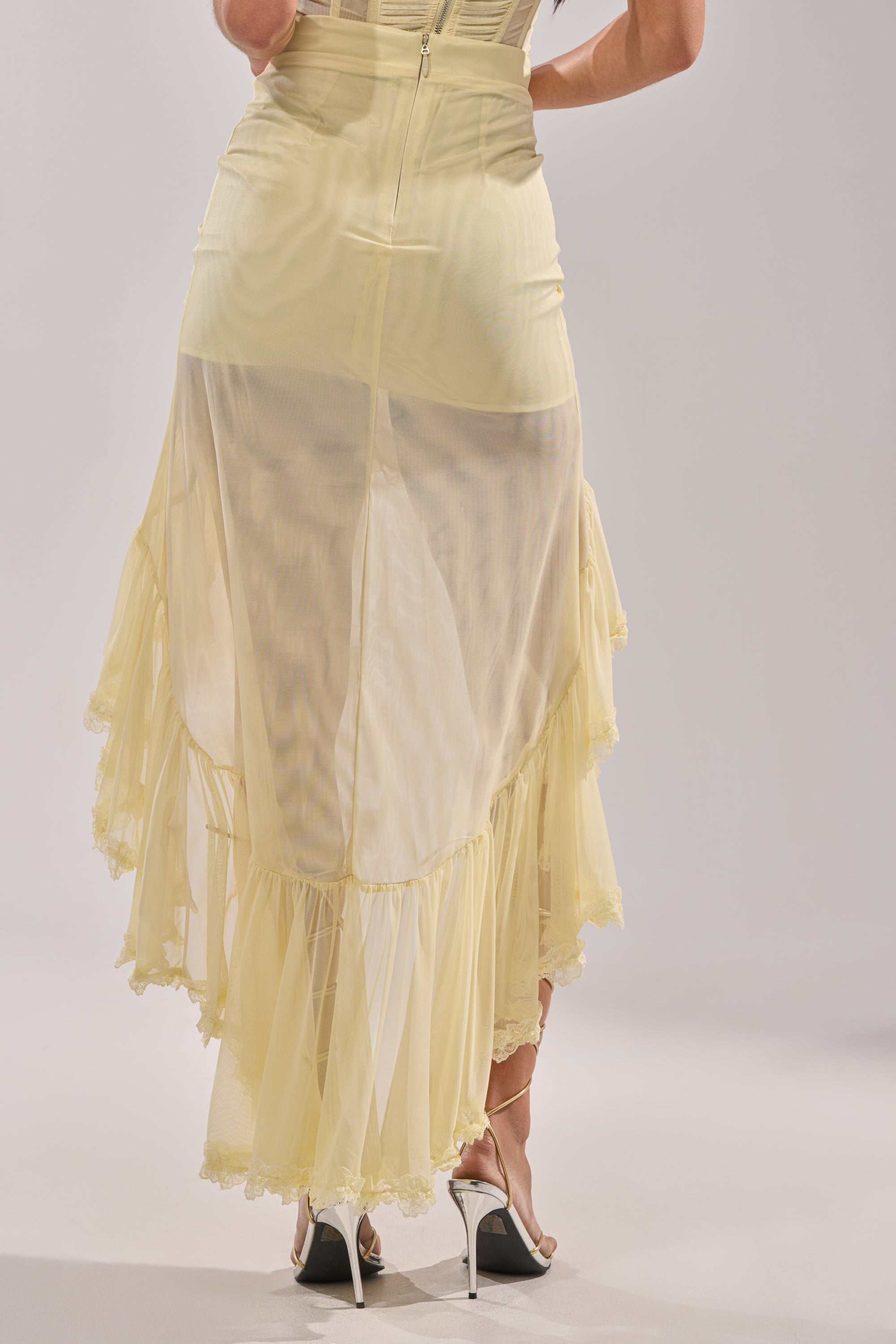 A person wearing a VACA VIBES MESH HIGH LOW SKIRT in pale yellow, paired with white high-heeled shoes, is shown from the back against a plain background. 7
