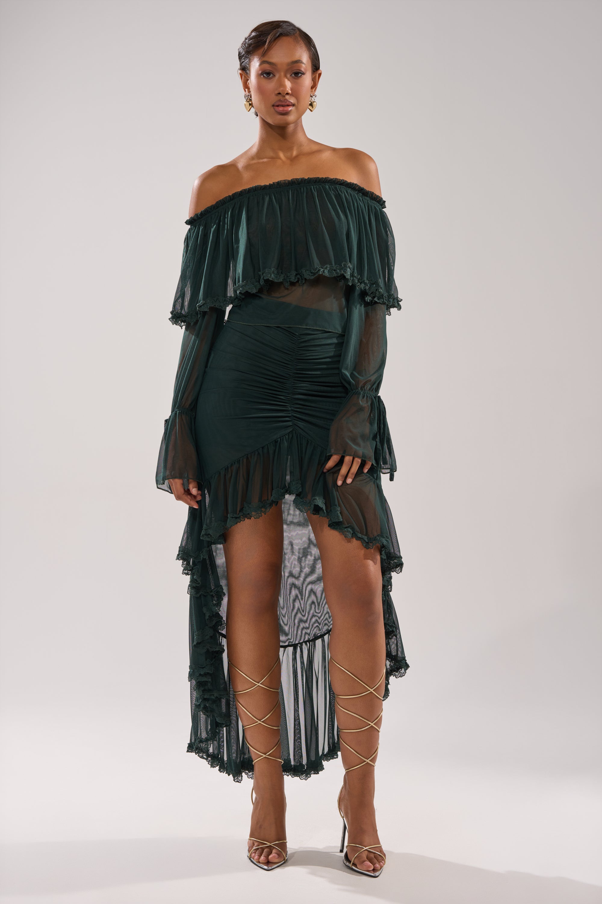 A woman poses against a plain light background, wearing the EMERALD RUCHED HIGH LOW SKIRT with sheer ruffled layers, styled with an off-shoulder top, strappy gold heels, and minimal jewelry. 0