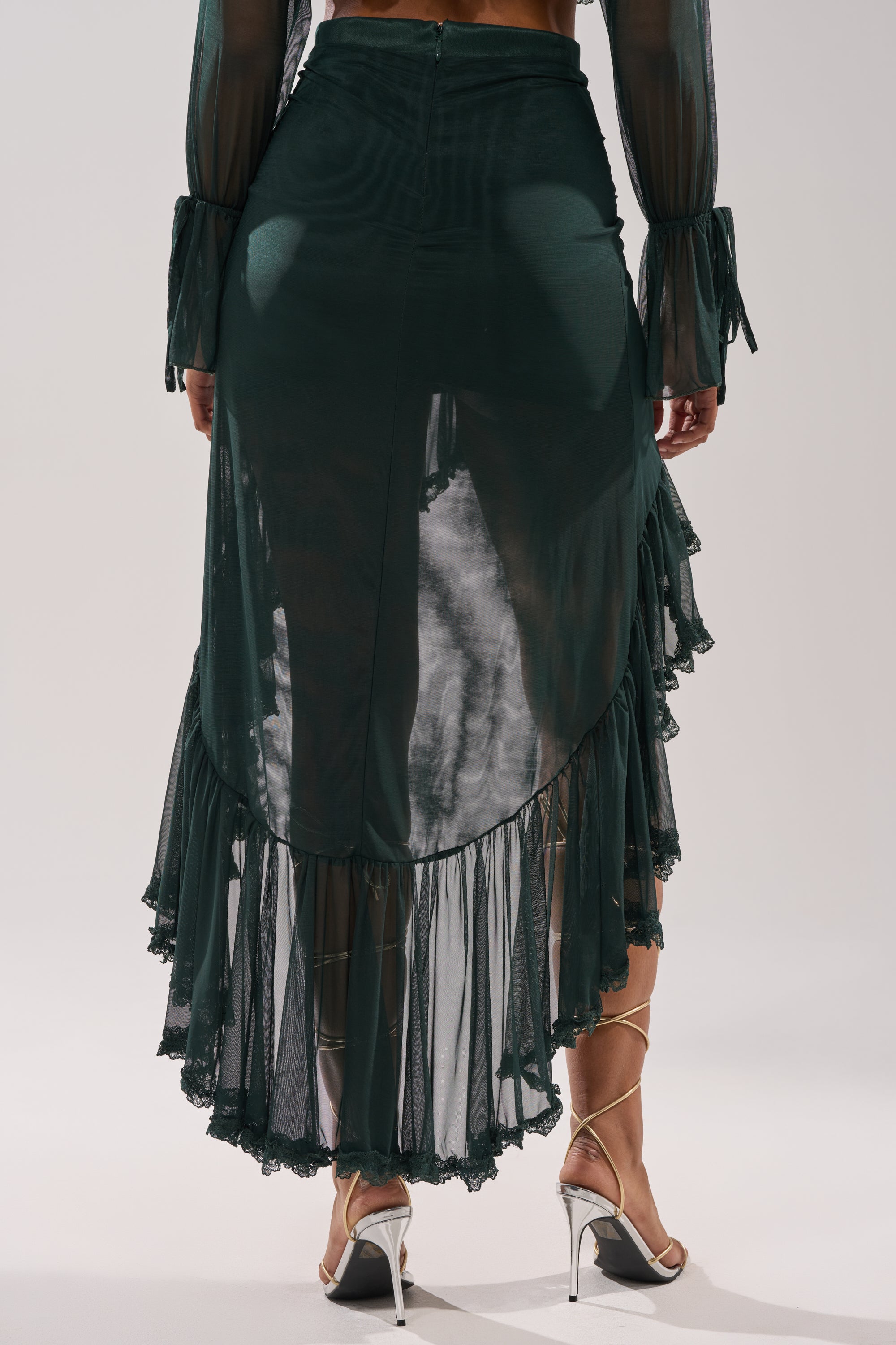 A person is shown from the back, wearing the EMERALD RUCHED HIGH LOW SKIRT with ruffle hem, paired with a matching top and silver high-heeled sandals, against a plain background. 3