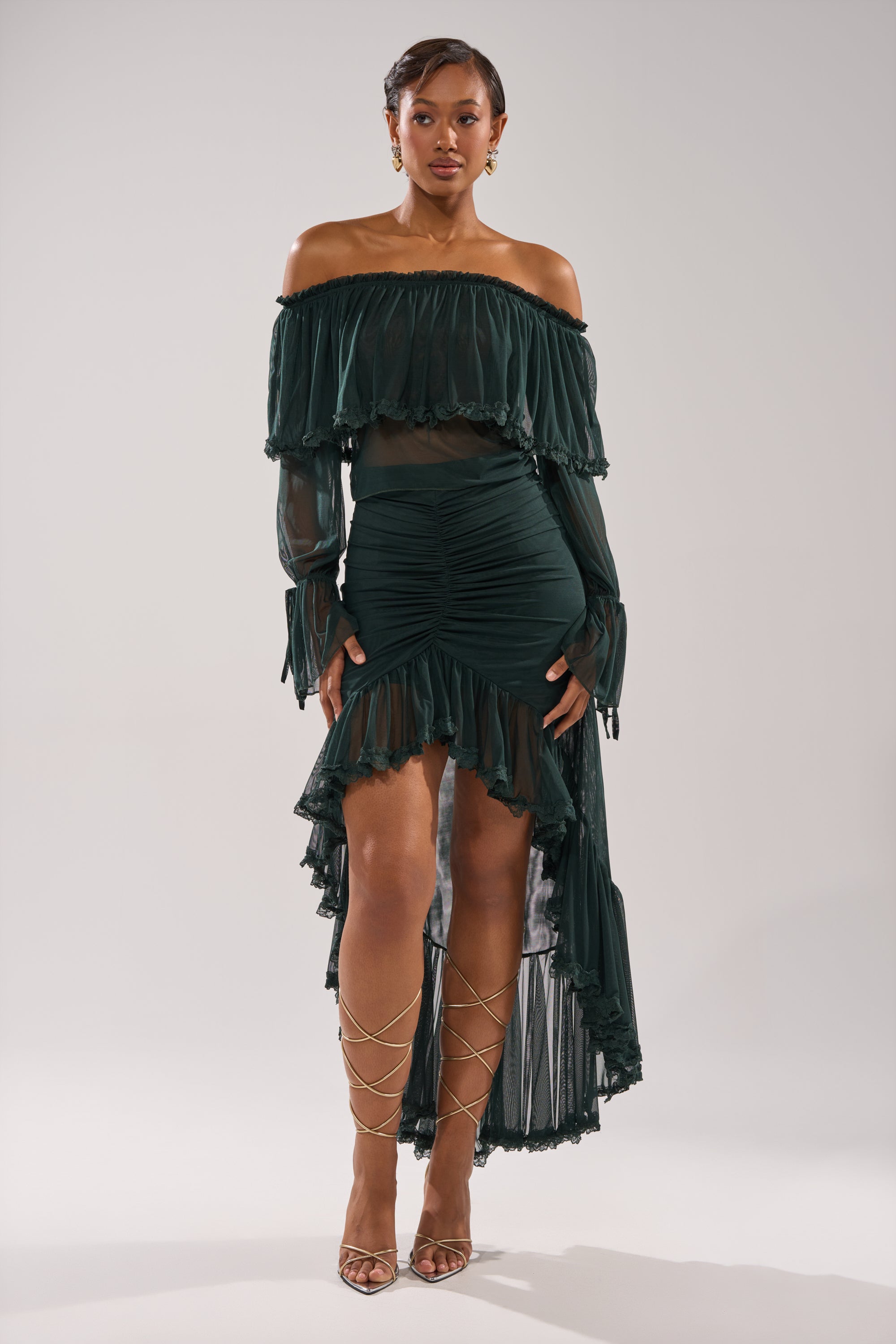 A woman faces forward in an off-the-shoulder dress paired with the EMERALD RUCHED HIGH LOW SKIRT, featuring a mesh ruffle and asymmetrical hem, styled with strappy gold heels against a plain light gray background. 4