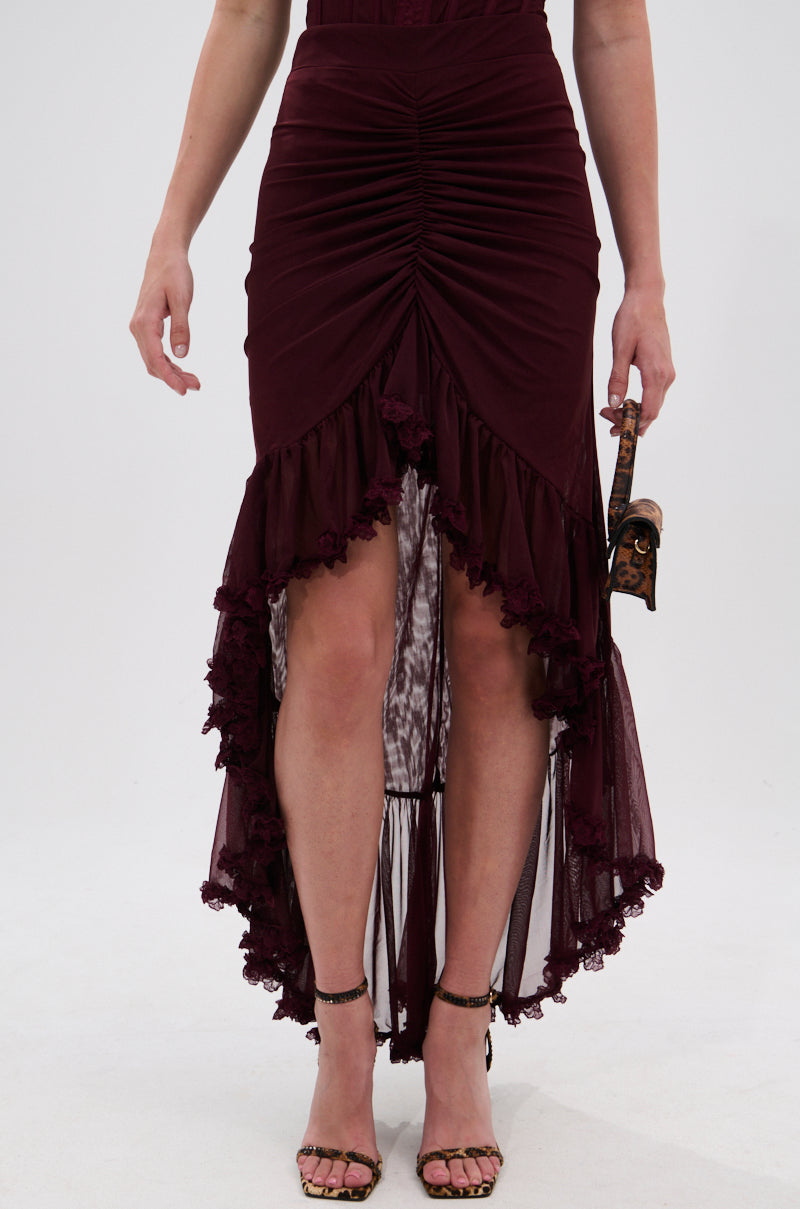 Someone wears the LOVELY DAYS MESH HIGH LOW SKIRT in burgundy, featuring a ruffled trim and ruched design, paired with strappy high-heeled sandals and carrying a small bag. 2