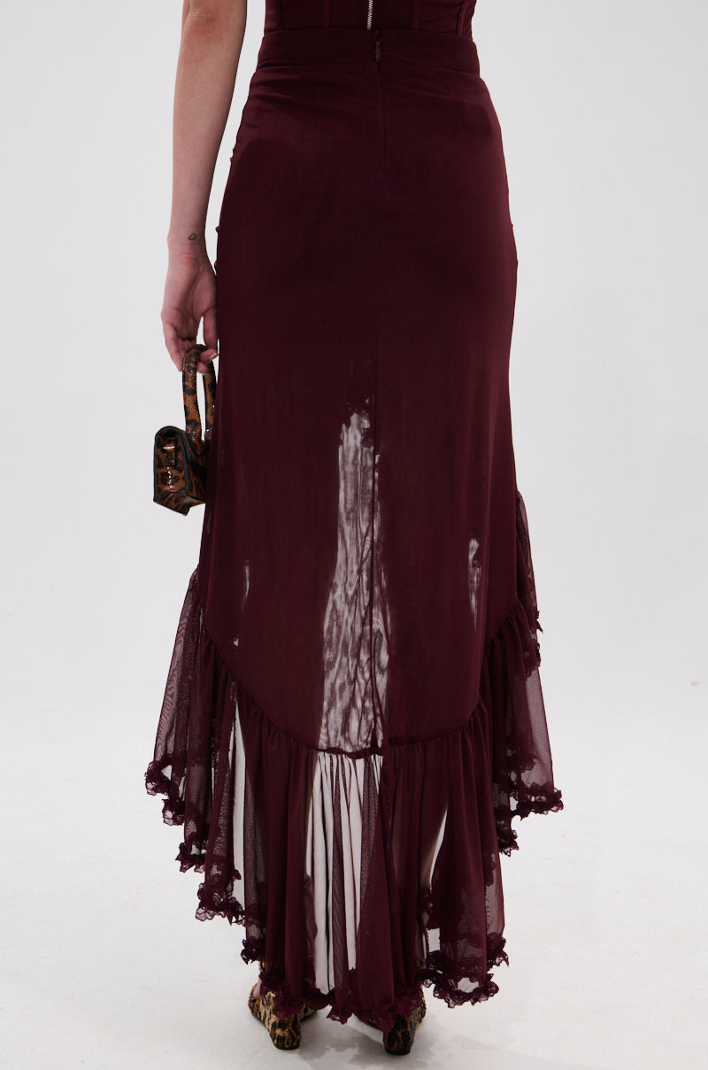 A person stands facing away, wearing the LOVELY DAYS MESH HIGH LOW SKIRT in burgundy with ruched details and lace-trimmed hem, holding a small handbag in their left hand against a plain white background. 4
