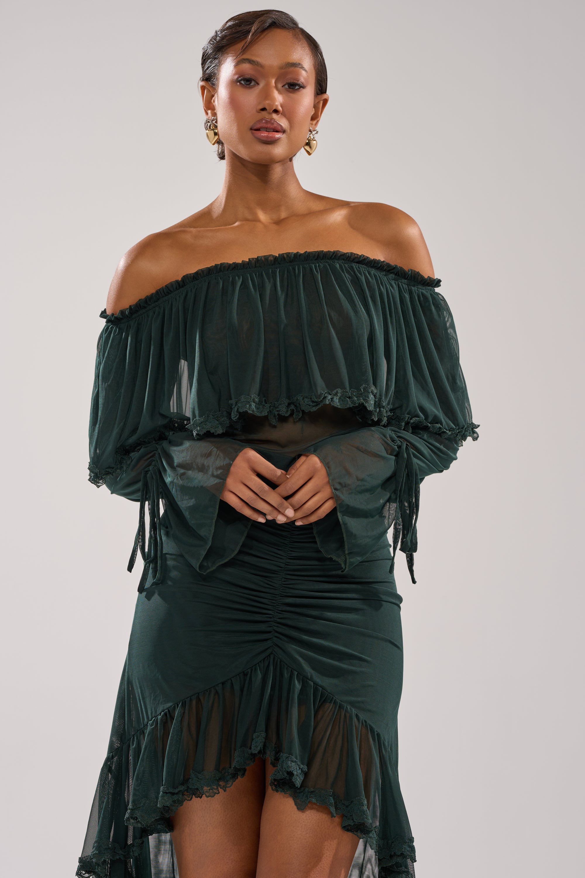 A woman confidently poses against a plain light background, wearing the EMERALD LOVER BLOUSE—an off-the-shoulder, dark green top with long sleeves and a ruched skirt.