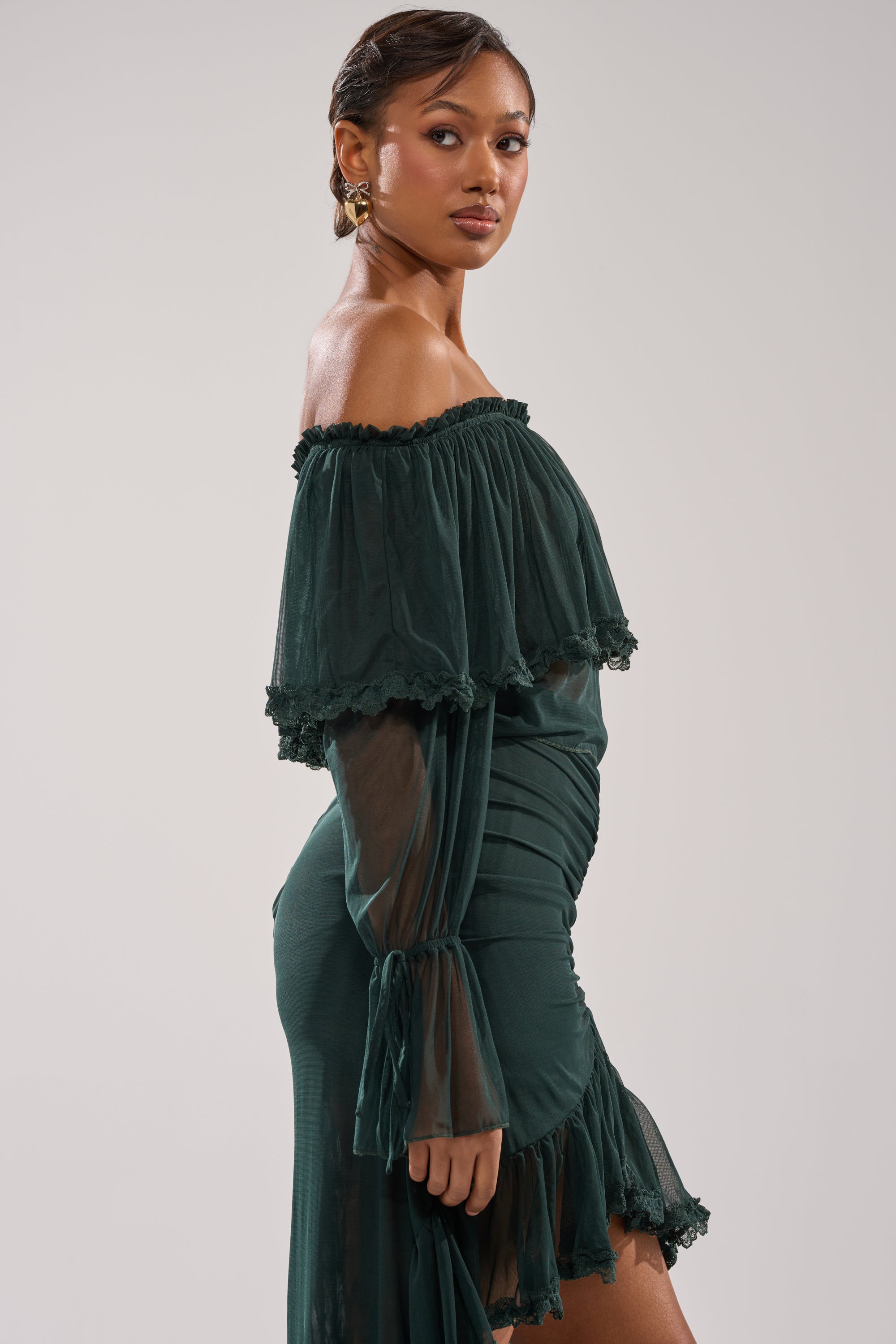 A woman in a dark green, off-the-shoulder EMERALD LOVER BLOUSE with ruffled trim stands in profile against a light background, her hair styled back as she gazes confidently at the camera.