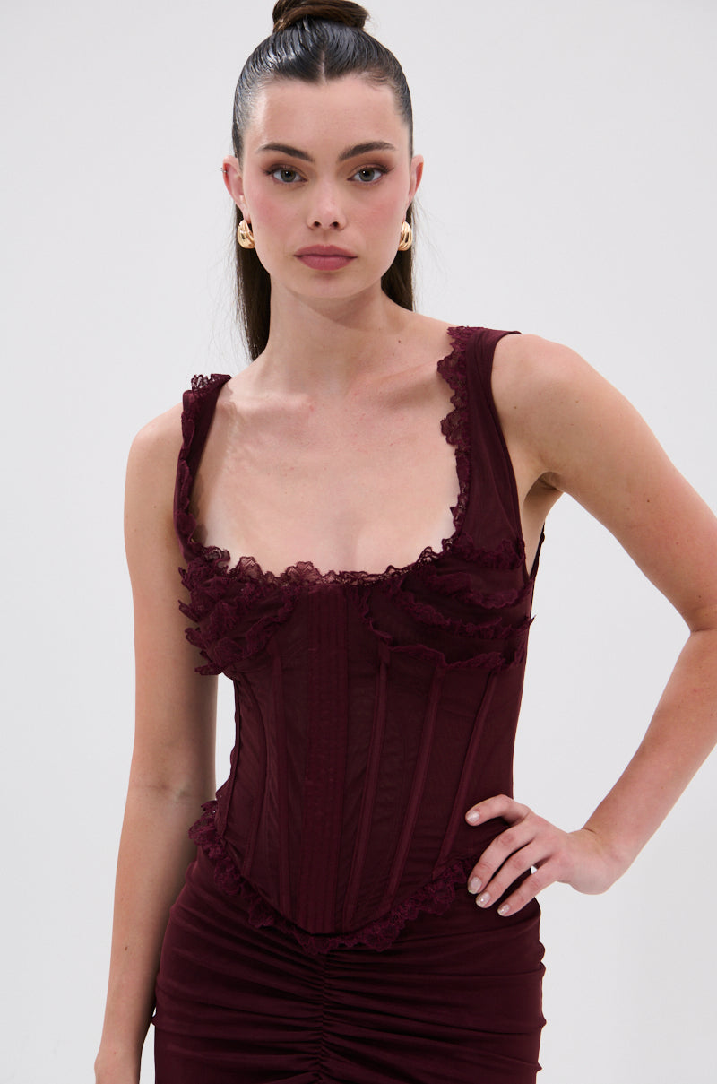 A woman with long dark hair in a sleek ponytail wears the LOVELY DAYS MESH CORSET, a deep burgundy mesh dress with ruffle lace trim. She poses confidently against a plain light background, one hand on her hip. 0