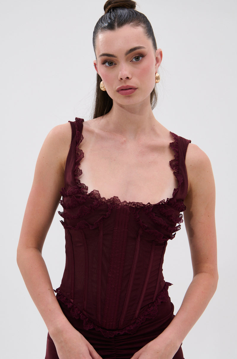 A woman with long brown hair in a high ponytail wears the LOVELY DAYS MESH CORSET, a sleeveless mesh top with ruffle lace trim. She styles it with gold hoop earrings and poses against a plain light background for a vintage look. 1