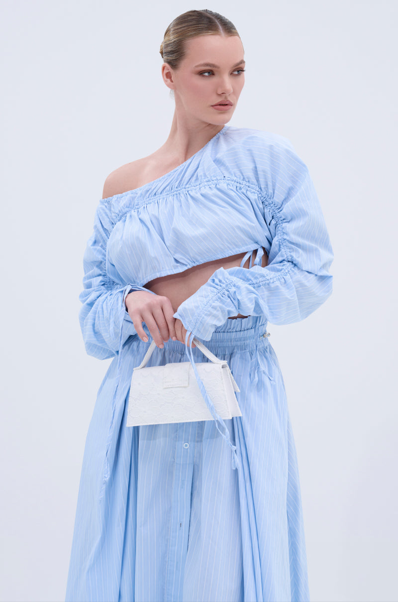A woman wears the TALK TO ME CINCHED BLOUSE in light blue with a matching flowy skirt, holding a small white handbag. Her hair is pulled back sleekly, and she poses against a light background.