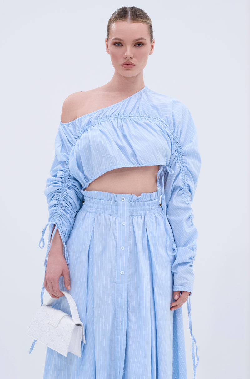 A woman poses in the TALK TO ME CINCHED BLOUSE, a chic light blue off-the-shoulder top with ruching, paired with a matching high-waisted button-down skirt. She carries a white handbag and stands against a plain light background.