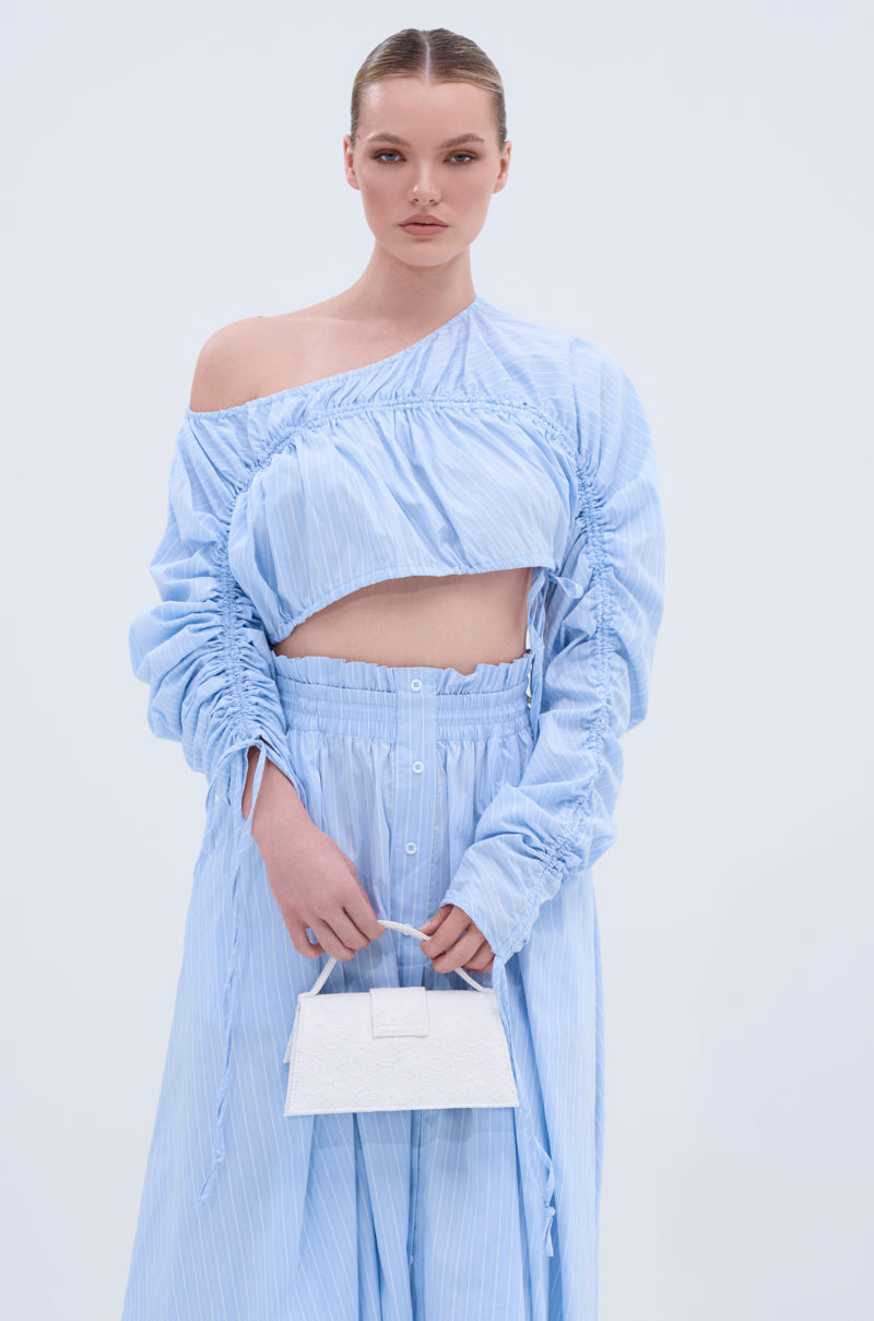 A woman wears the TALK TO ME CINCHED BLOUSE in light blue, styled off-the-shoulder with a matching high-waisted skirt and a small white handbag, standing against a plain light background, looking at the camera.