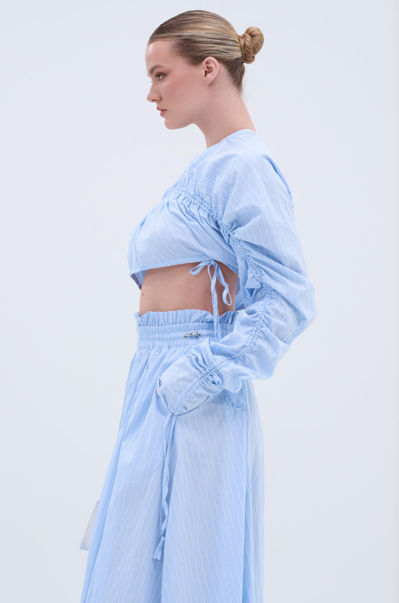 A woman with blonde hair in a neat bun models the TALK TO ME CINCHED BLOUSE, a light blue top with tie details, paired with a matching high-waisted skirt. She is shown in profile against a plain light background.
