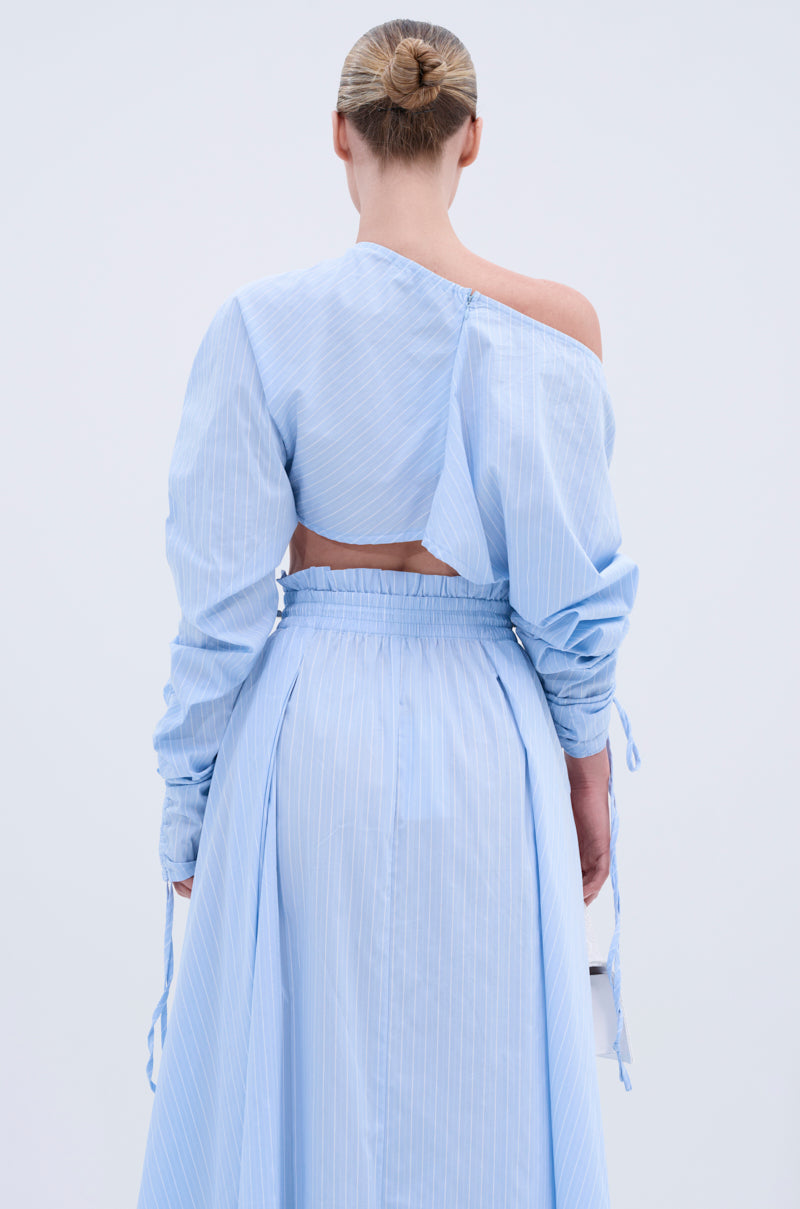 A woman with blonde hair in a bun is seen from behind wearing the TALK TO ME CINCHED BLOUSE, a light blue pinstriped cotton top with one-shoulder, long sleeves, and an exposed back detail for added elegance.