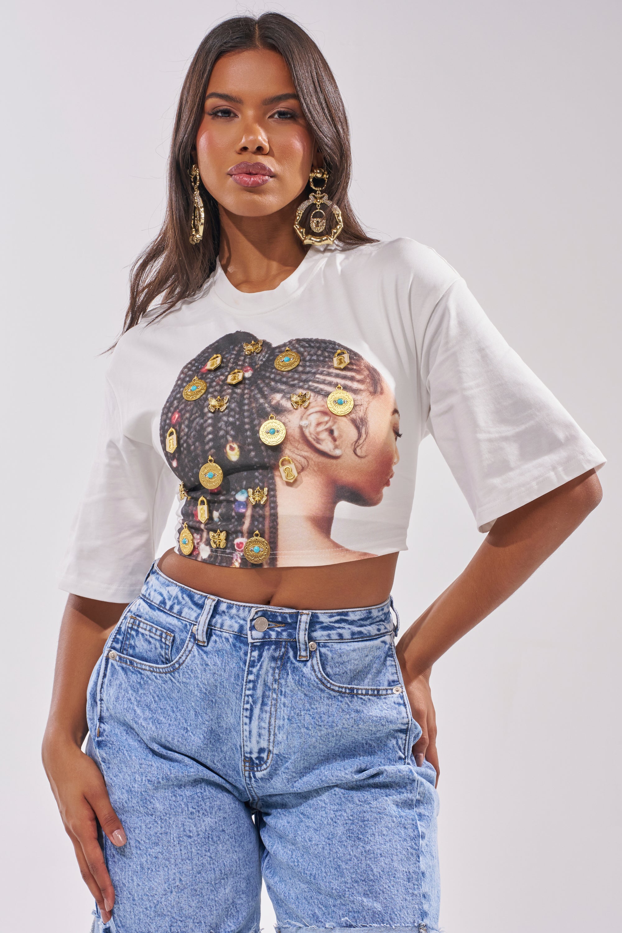 A woman wears the LOCKED AND LOADED EMBELLISHED TEE, a cropped graphic top with a beaded braids design, paired with high-waisted blue jeans—her bold look and gold earrings make this outfit perfect for standout festival wear. 0