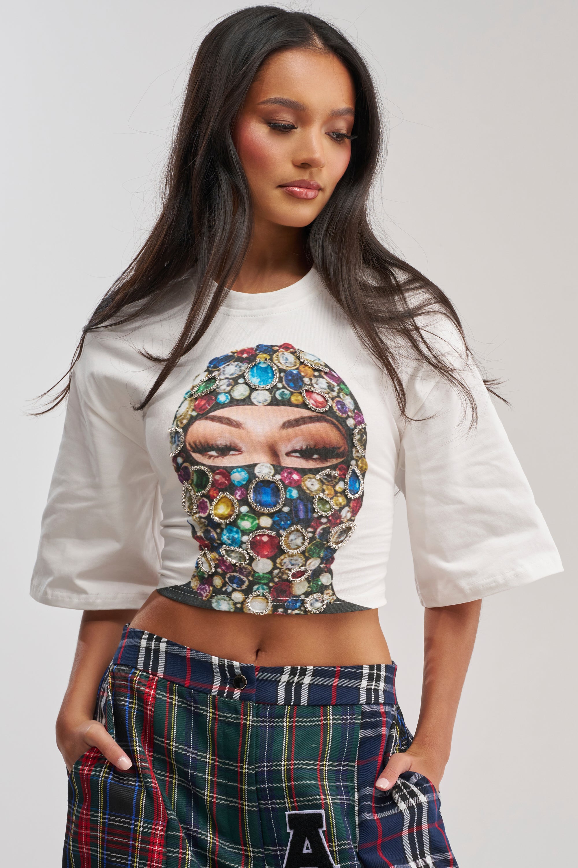 A woman with long dark hair models the FACE CARD GRAPHIC CROPPED TEE, which features a jeweled ski mask graphic. She pairs it with plaid pants, hands in pockets, and poses against a plain background for an effortless streetwear look.