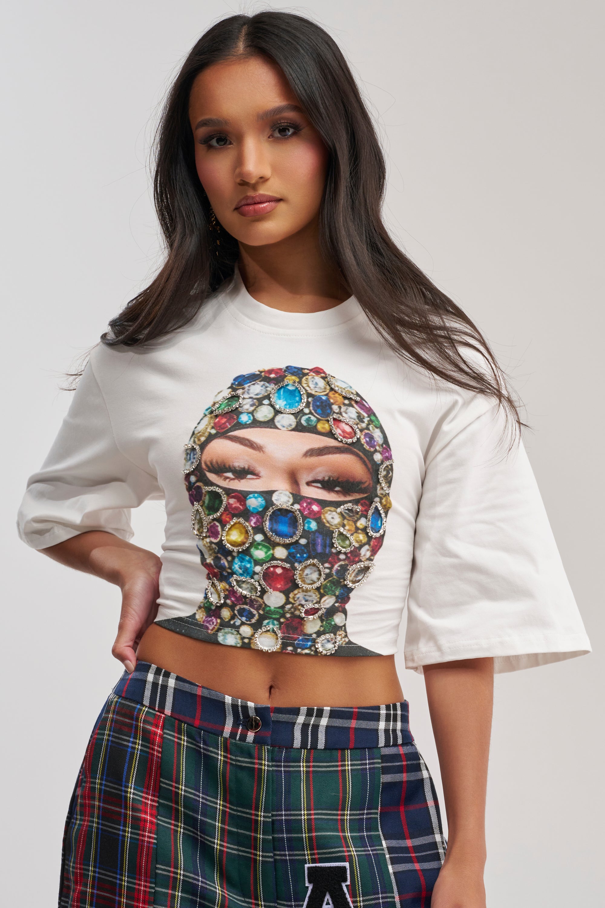 A woman with long dark hair models the FACE CARD GRAPHIC CROPPED TEE, showcasing a jeweled mask graphic with rhinestone chain accents. She pairs it with plaid pants and poses against a plain light background, one hand on her hip.