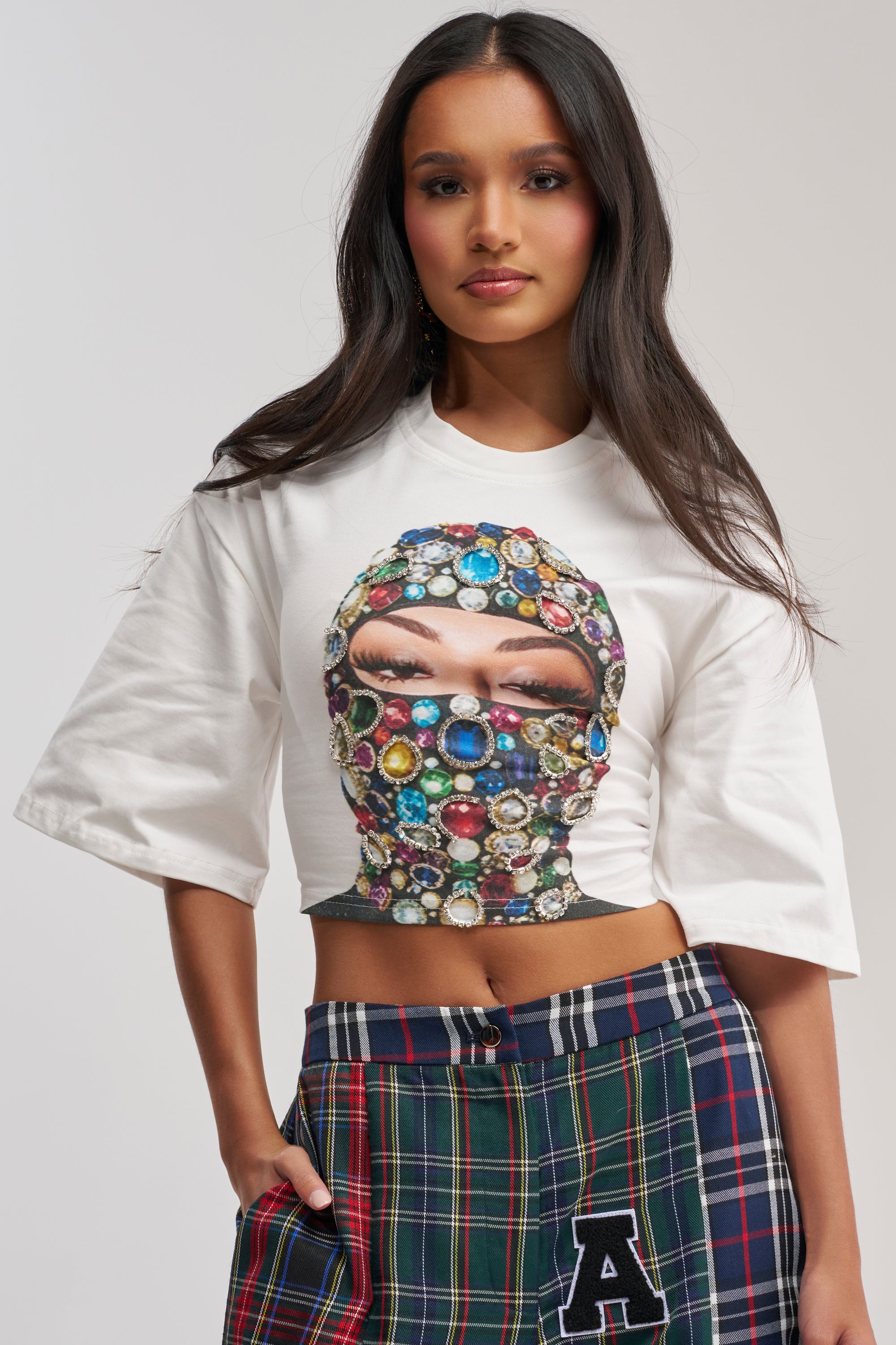 A woman with long dark hair wears the FACE CARD GRAPHIC CROPPED TEE, featuring a jeweled mask graphic, paired with plaid streetwear pants with an "A" patch, posing against a neutral background.