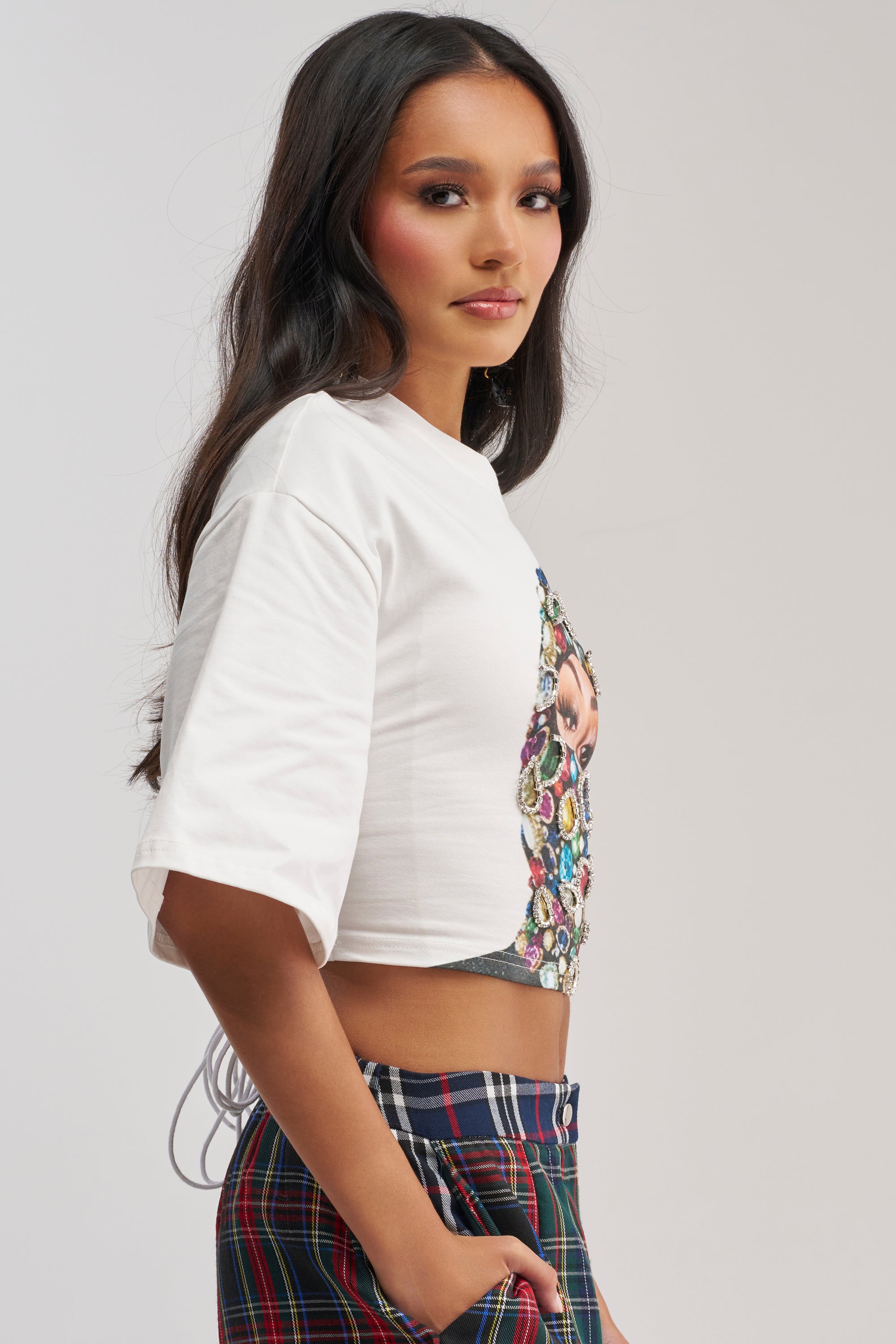 A woman with long dark hair models the FACE CARD GRAPHIC CROPPED TEE and plaid pants, standing sideways against a plain light background and looking at the camera.